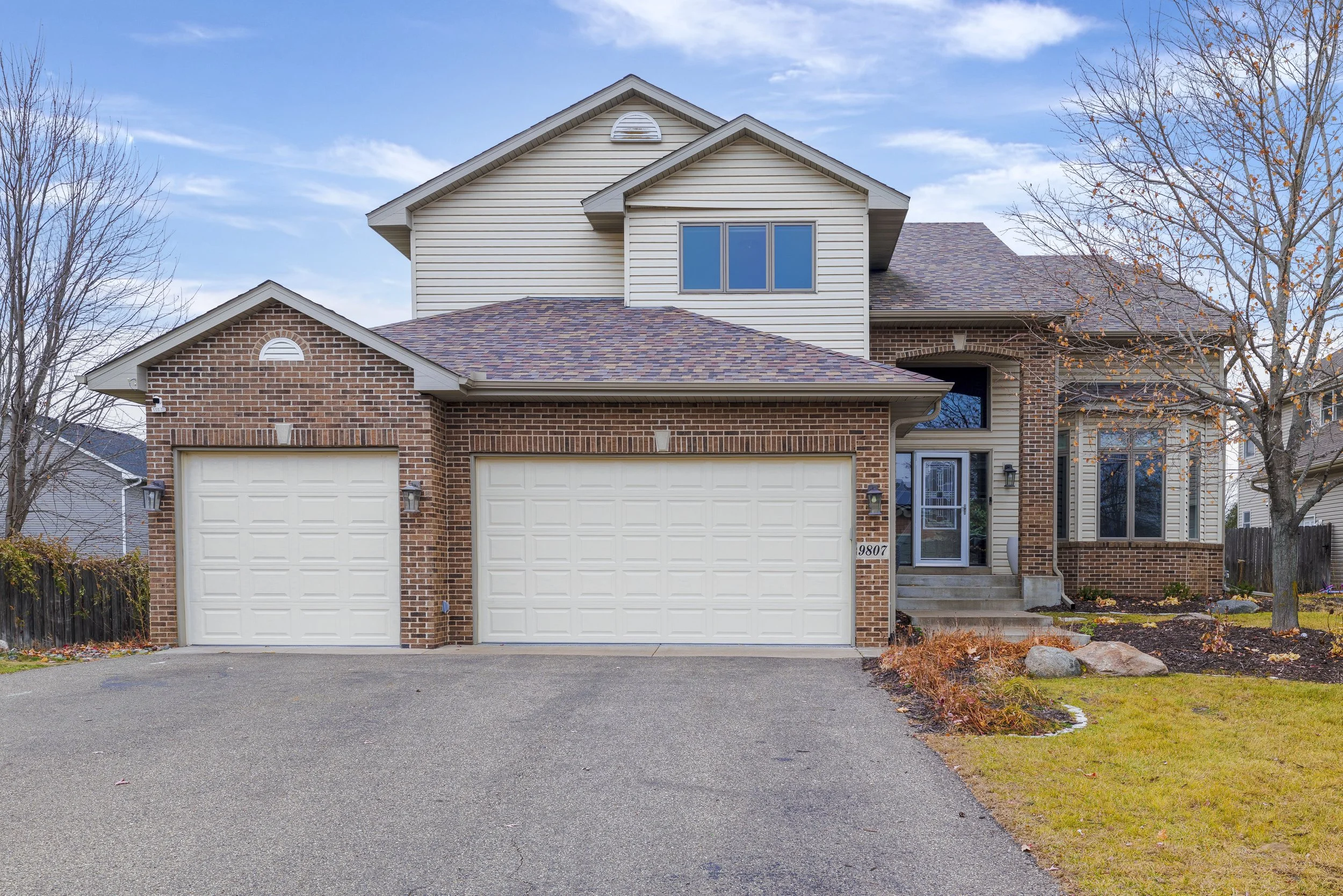 Front view of a two-story house with brick and beige siding, two white garage doors, a paved driveway, leafless trees, and a manicured lawn.