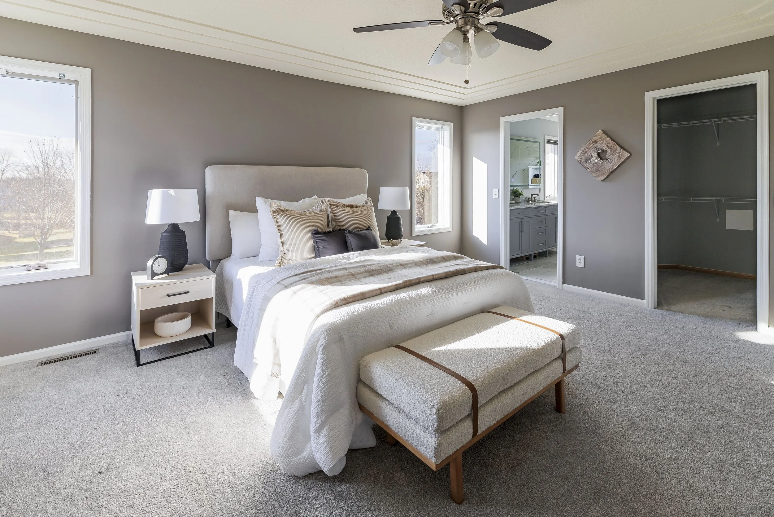 A bedroom with a bed, nightstands, lamps, and a window overlooking an outdoor yard. There is a bench at the foot of the bed, an open closet, and a view into the bathroom.