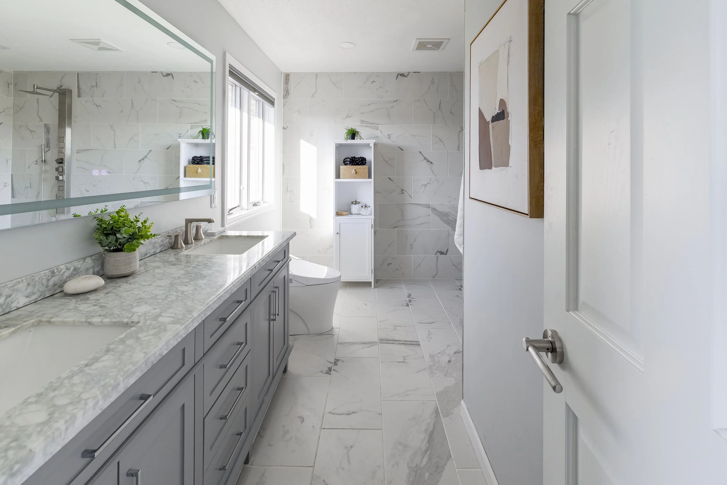 Modern bathroom with marble countertops, gray cabinetry, large mirror, shower area with glass door, and white toilet, decorated with plants and artwork.
