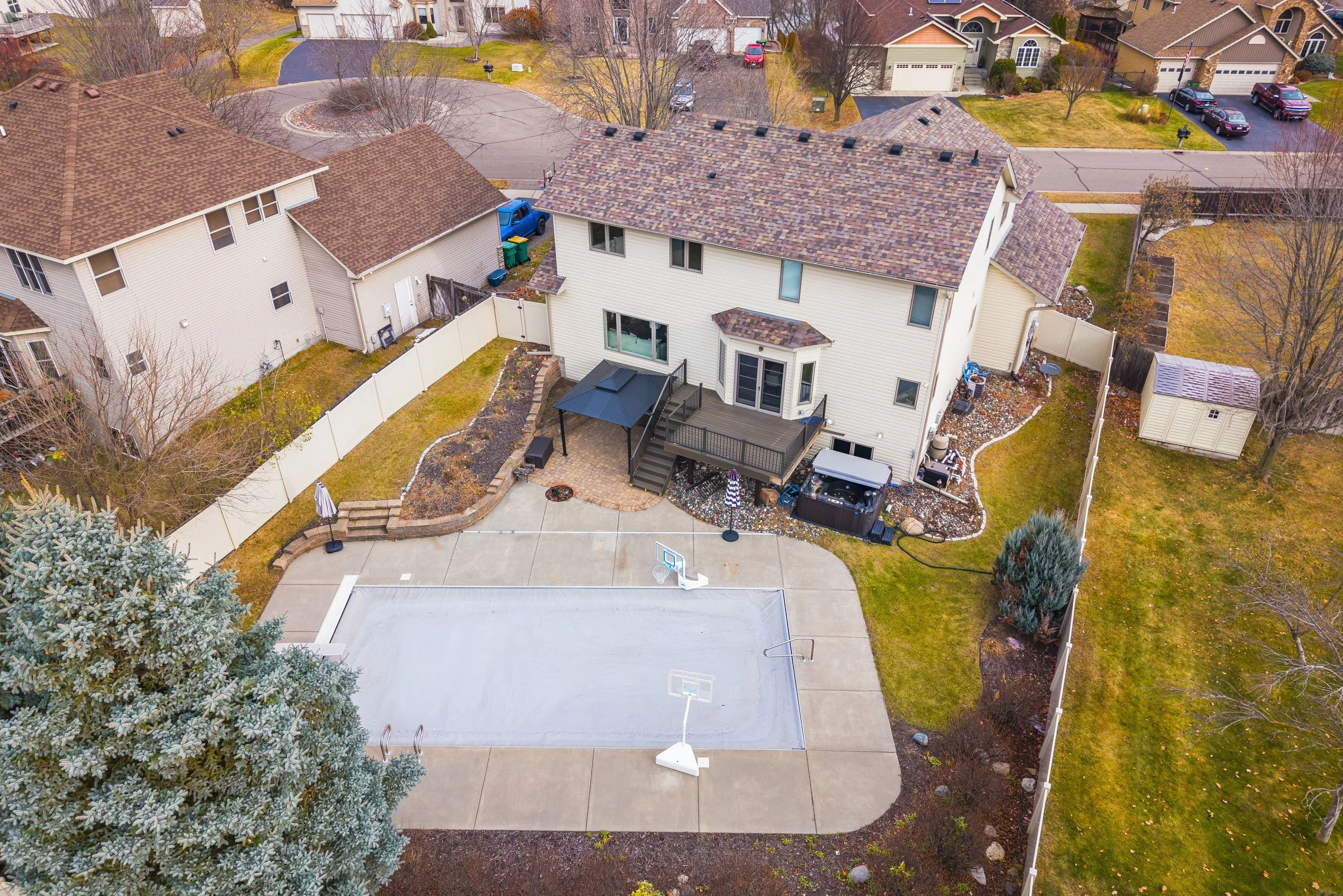 A backyard with a concrete basketball court and two basketball hoops, a multi-level deck with stairs, a canopy, and a hot tub. The yard is fenced and surrounded by neighboring houses, trees, and grassy areas.