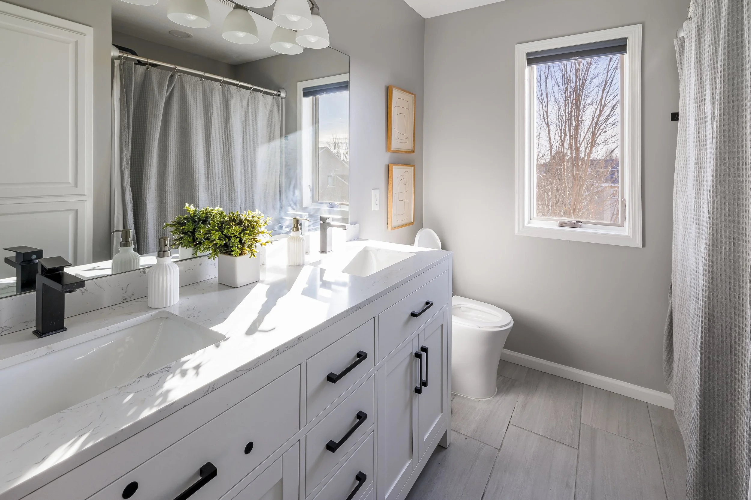 Modern bathroom with white double vanity, black handles, marble countertop, two sinks, and a toilet. There is a large mirror, window with blinds, and a shower with a curtain. Decor includes a potted plant and soap dispensers.