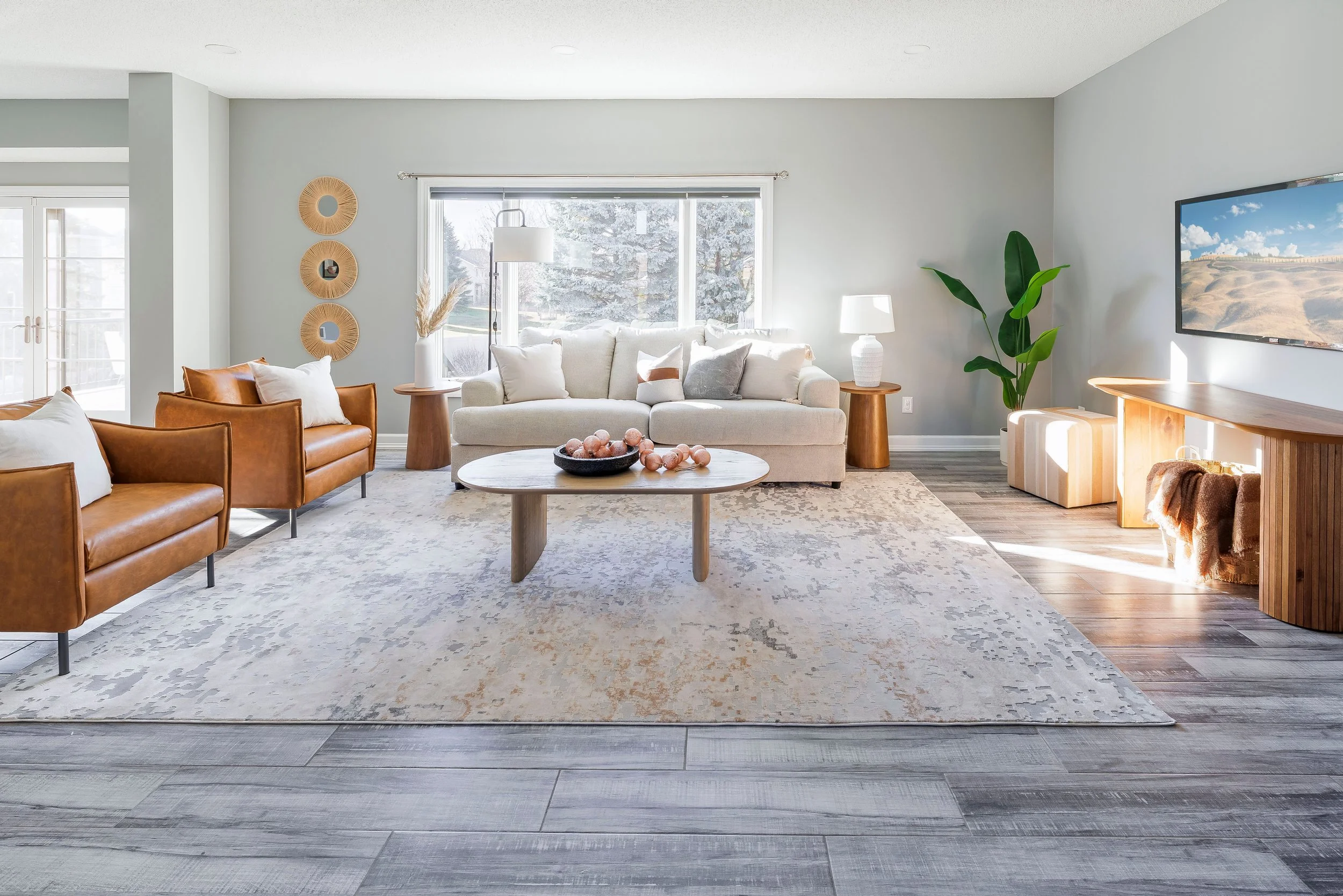 Bright living room with large window, white couch, two wooden side tables with lamps, two leather armchairs, a round coffee table with decor, a large patterned rug, a TV on a wooden stand, and a potted plant.