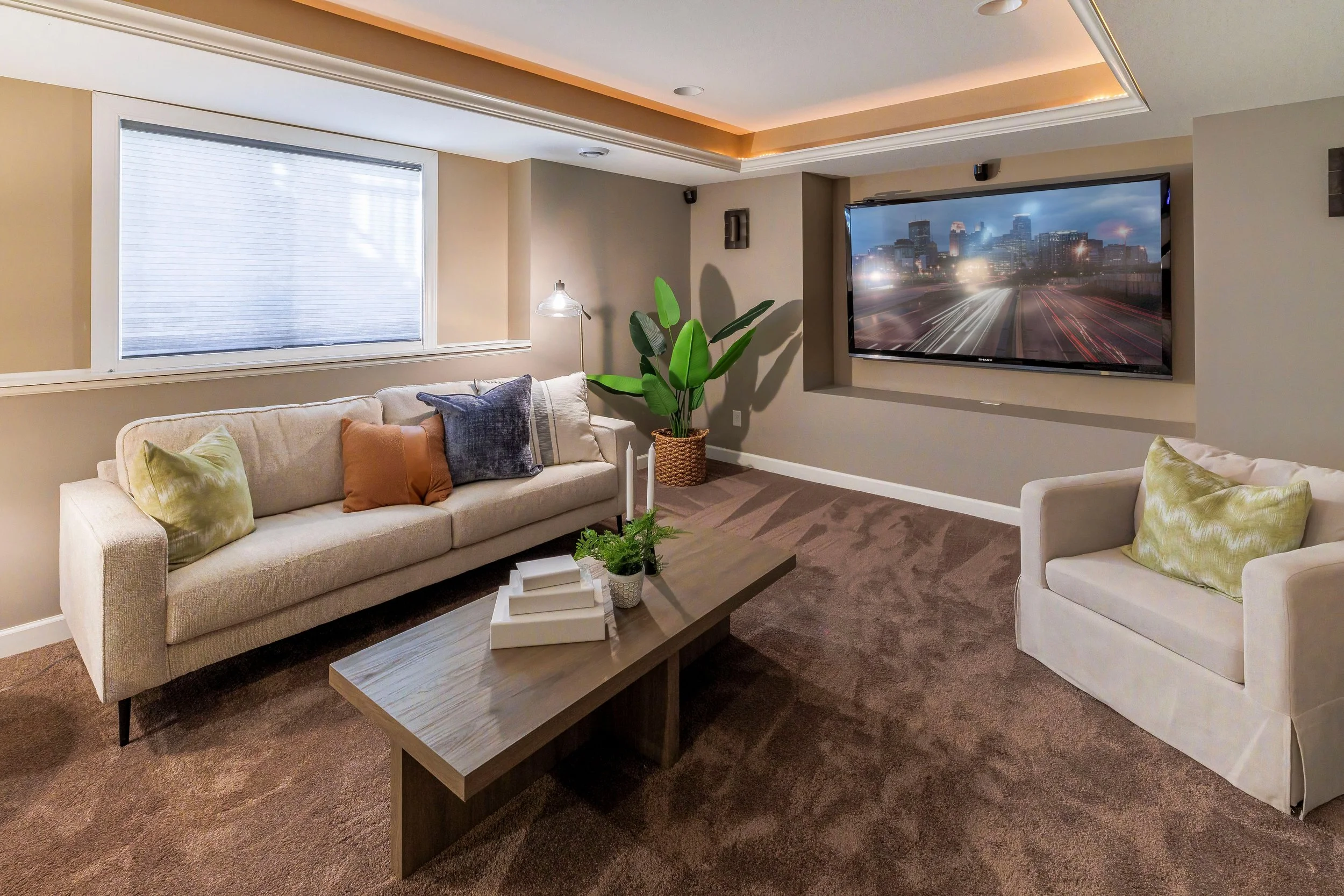 Living room with beige couches, decorative pillows, a wooden coffee table with books and a plant, a large window with closed blinds, a potted plant, a mounted flat-screen TV showing a cityscape with moving traffic, and soft ceiling lighting.