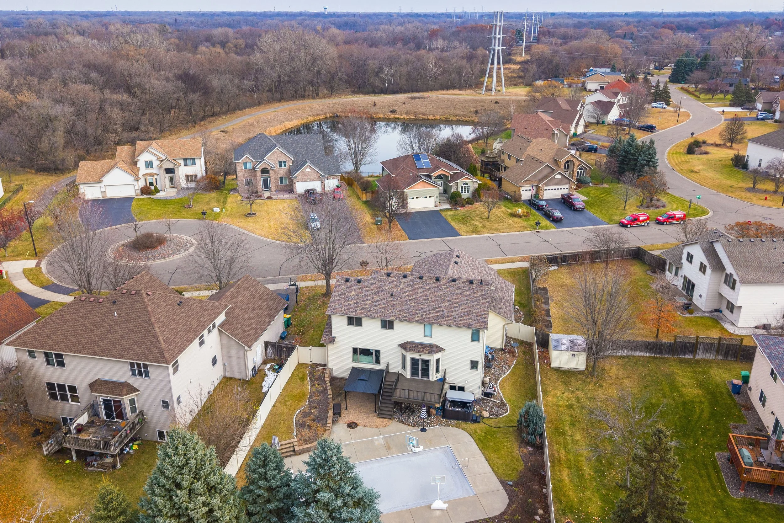 Aerial view of a suburban neighborhood with houses, a pond, and a basketball court, surrounded by trees and paved streets.
