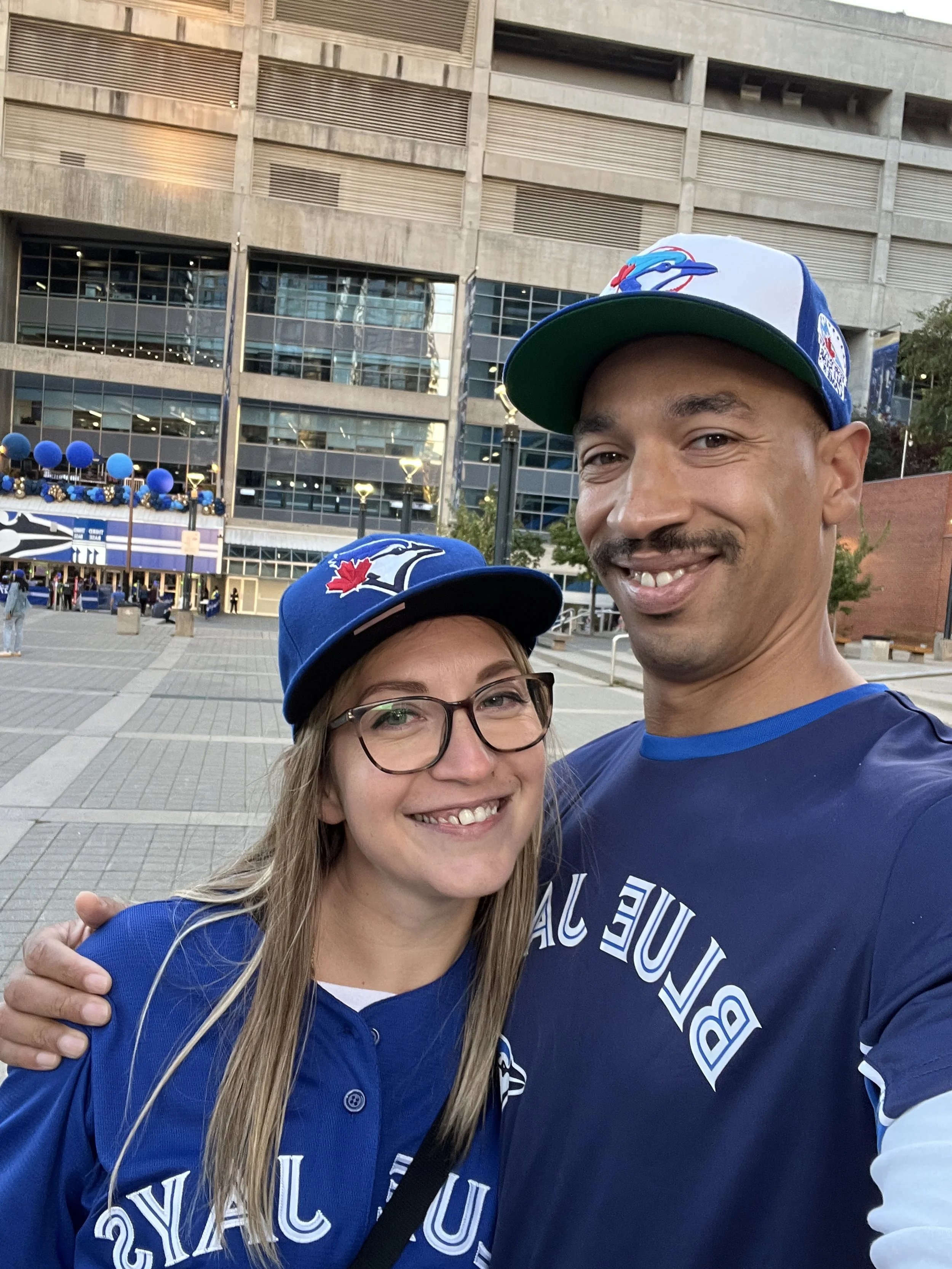 A smiling man and woman taking a selfie outside a stadium, both wearing blue Toronto Blue Jays hats and jerseys, with the stadium and crowd in the background.