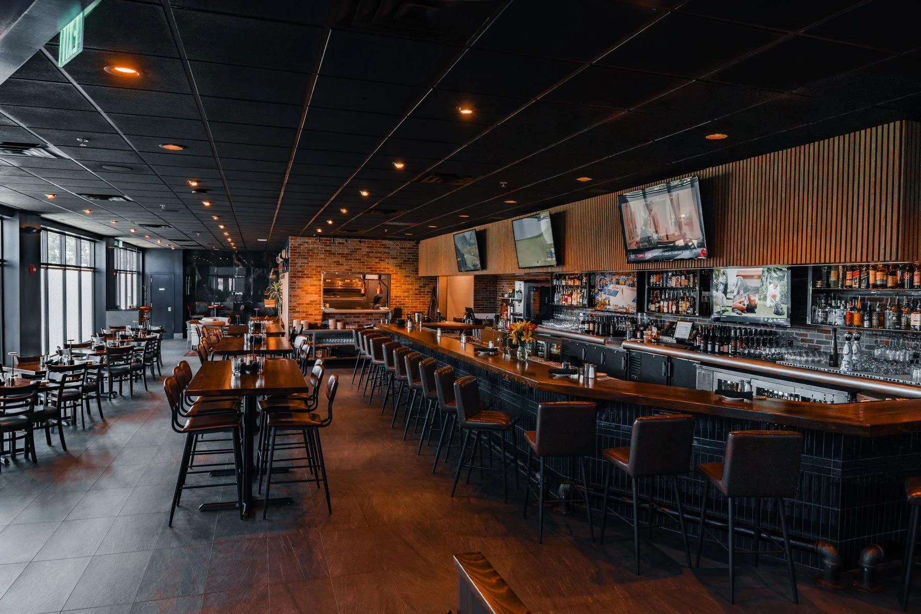 Interior of a modern bar and restaurant with dark ceilings, large windows, a bar with high chairs, multiple TVs mounted on a wooden-paneled wall, and tables set for dining.