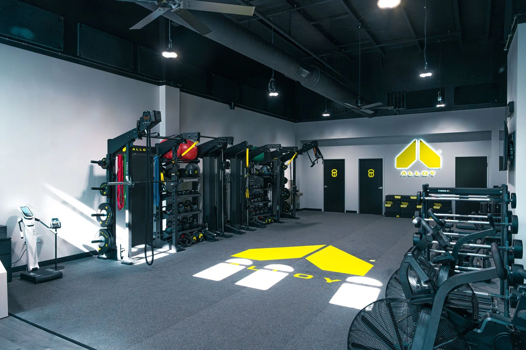 Interior of a modern gym with workout equipment, weight racks, and a illuminated yellow and white logo on the wall that says "ALLOY."