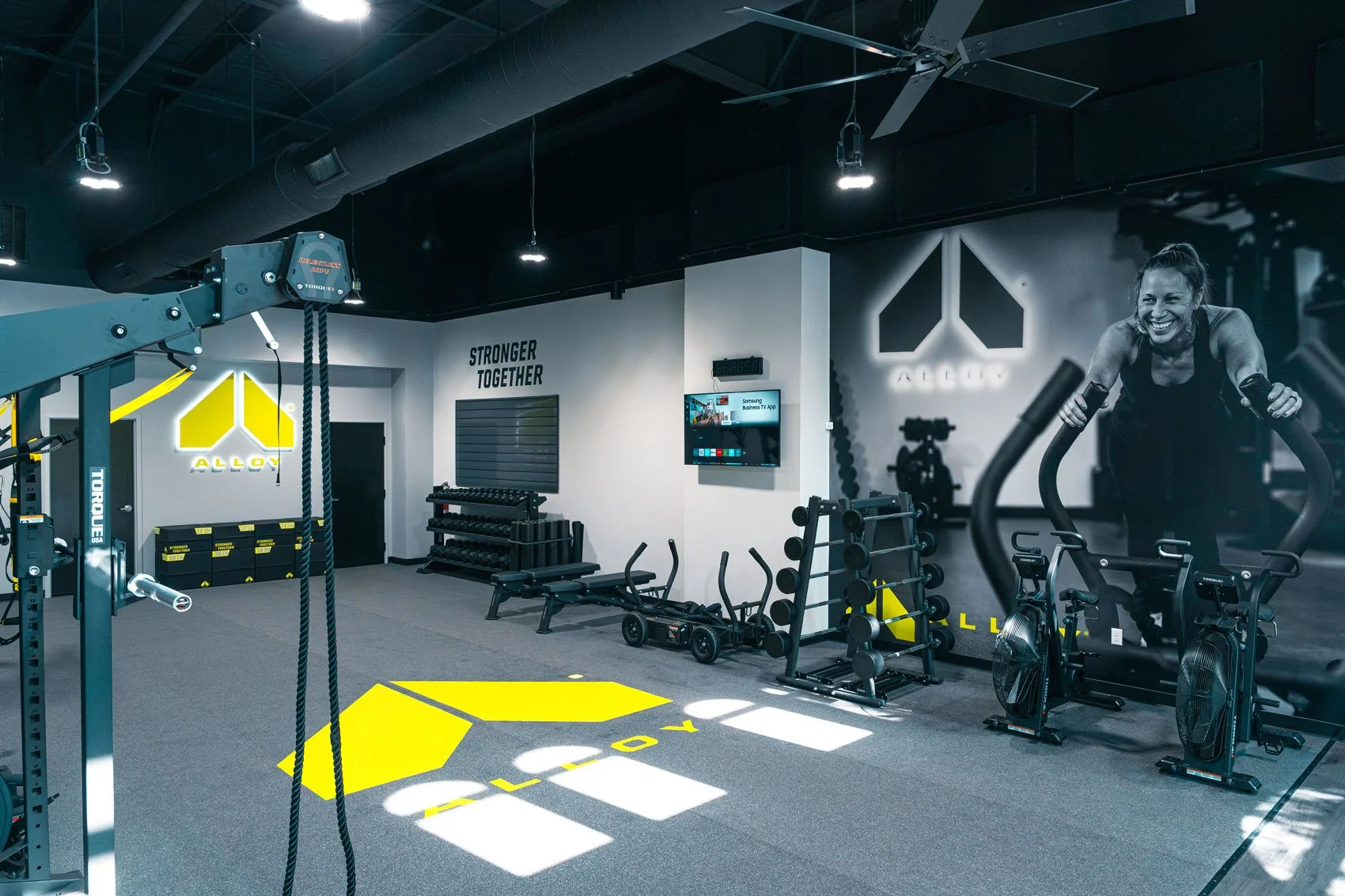 Modern gym interior with workout equipment, large wall graphic of a smiling woman exercising, and illuminated 'ALLOY' logo on wall