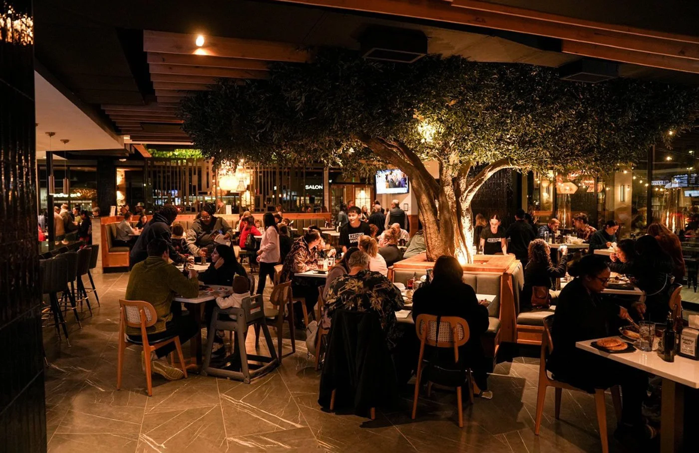 Interior of a restaurant with a large central tree, people dining at tables, warm ambient lighting, and modern decor.