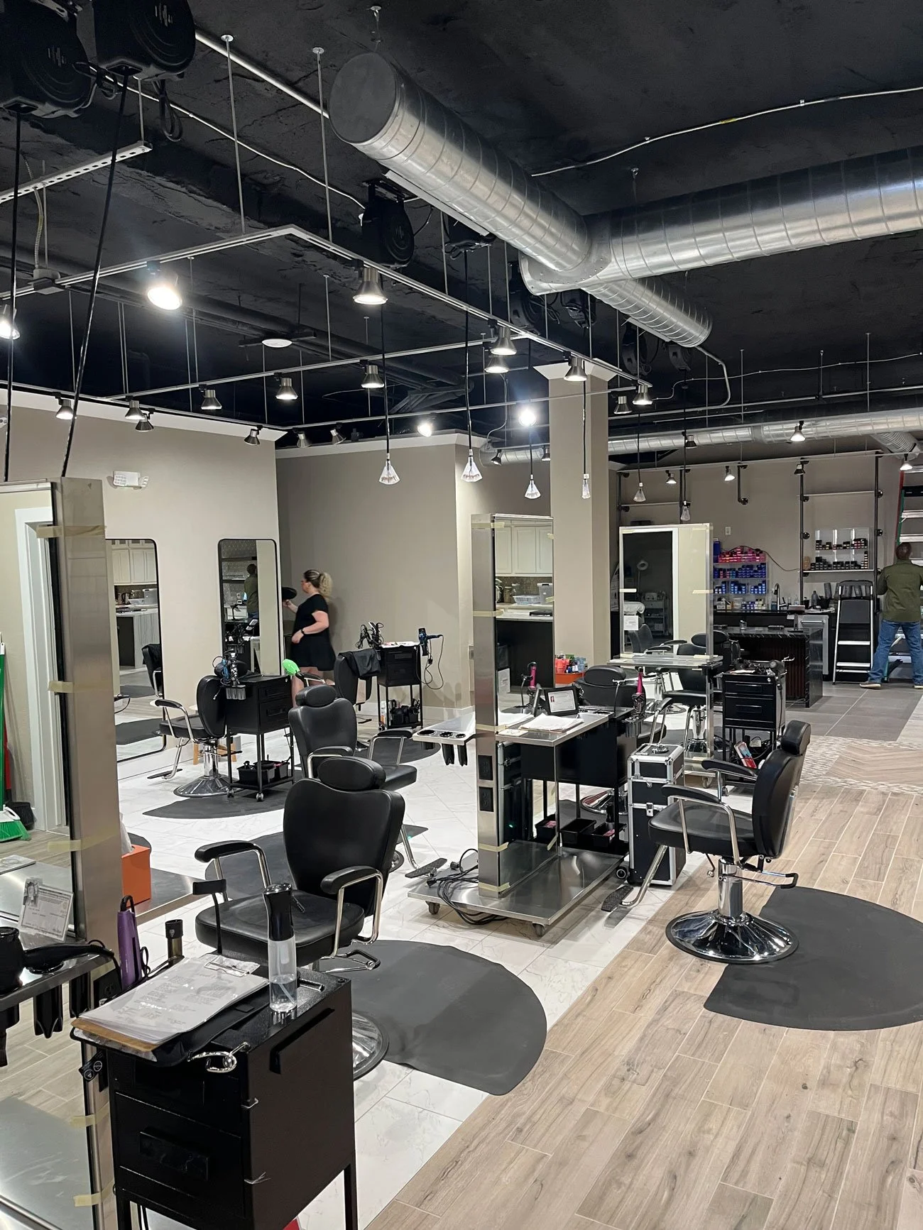 Interior of a modern hair salon with black chairs, mirrors, and hair styling stations, some people working and walking inside.