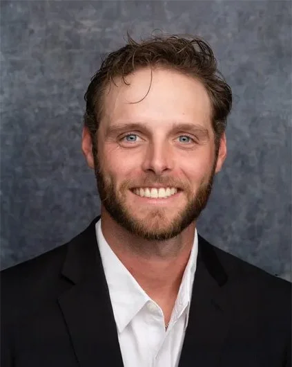 Professional headshot of a man with light brown hair, blue eyes, beard, wearing a black suit jacket and white shirt, smiling against a gray textured background.