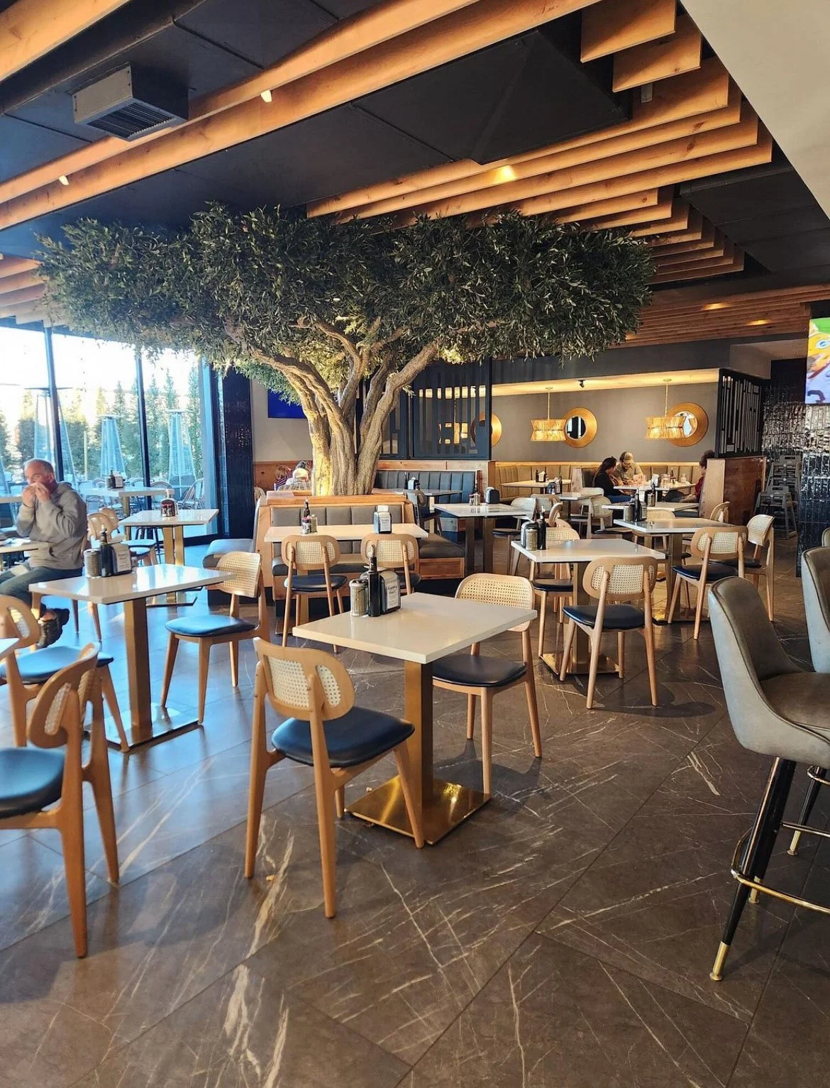 Restaurant interior with large artificial tree, modern decor, and seating area with tables and chairs, some people dining.
