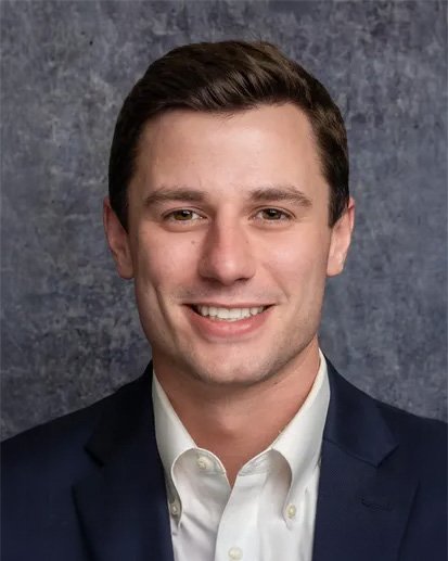 A professional headshot of a young man with dark hair in a suit, smiling in front of a textured gray background.