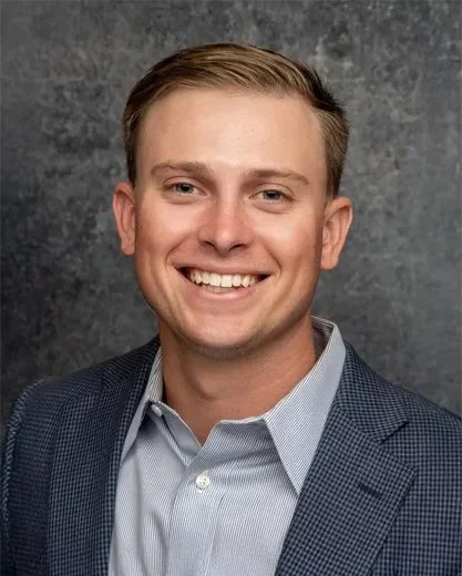 Portrait of a young man with short light brown hair, smiling, wearing a light blue dress shirt and a dark blue blazer, against a textured gray background.
