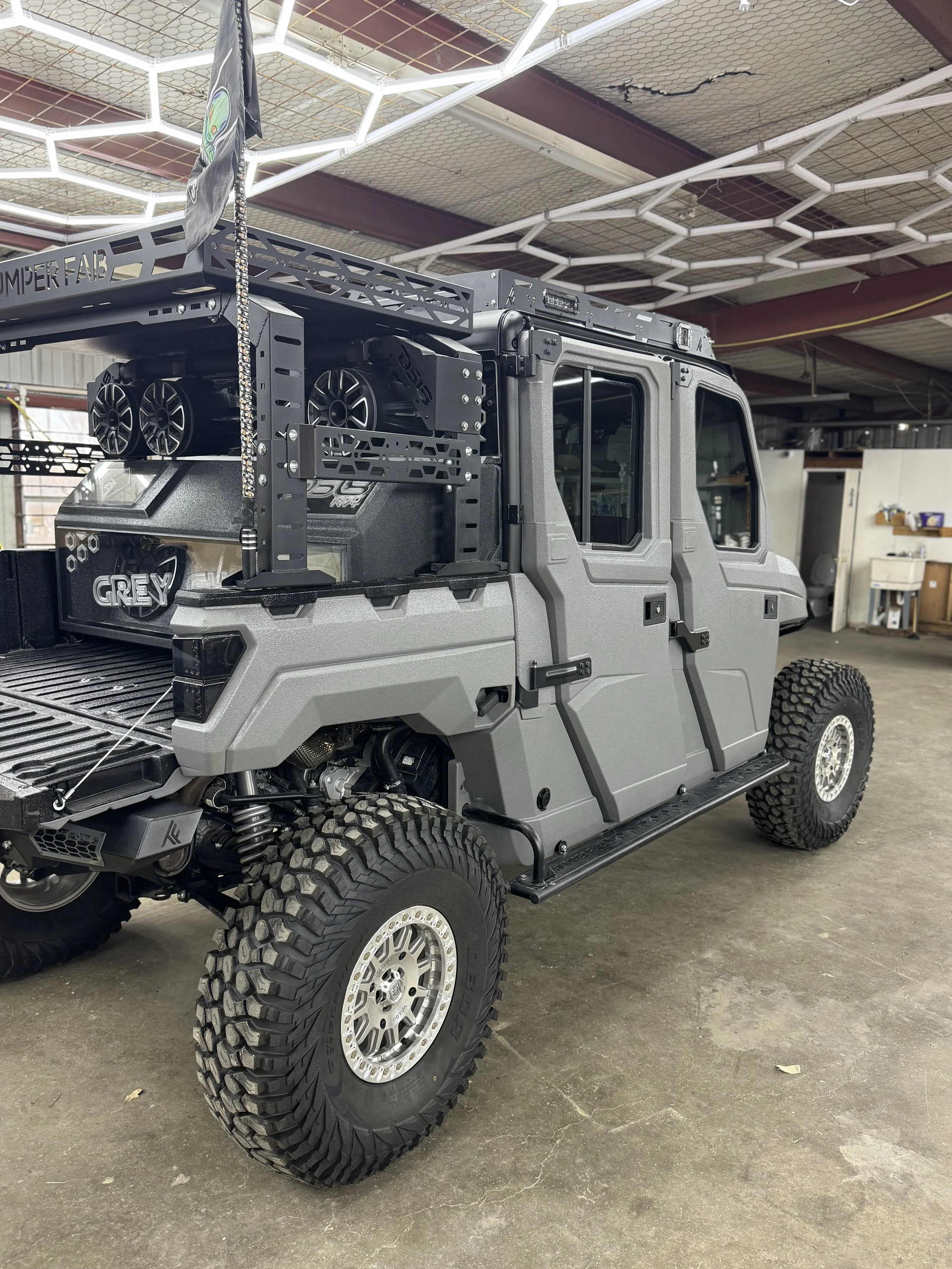 A customized gray off-road utility vehicle with large rugged tires, robust suspension, and a roof rack, parked inside a warehouse.