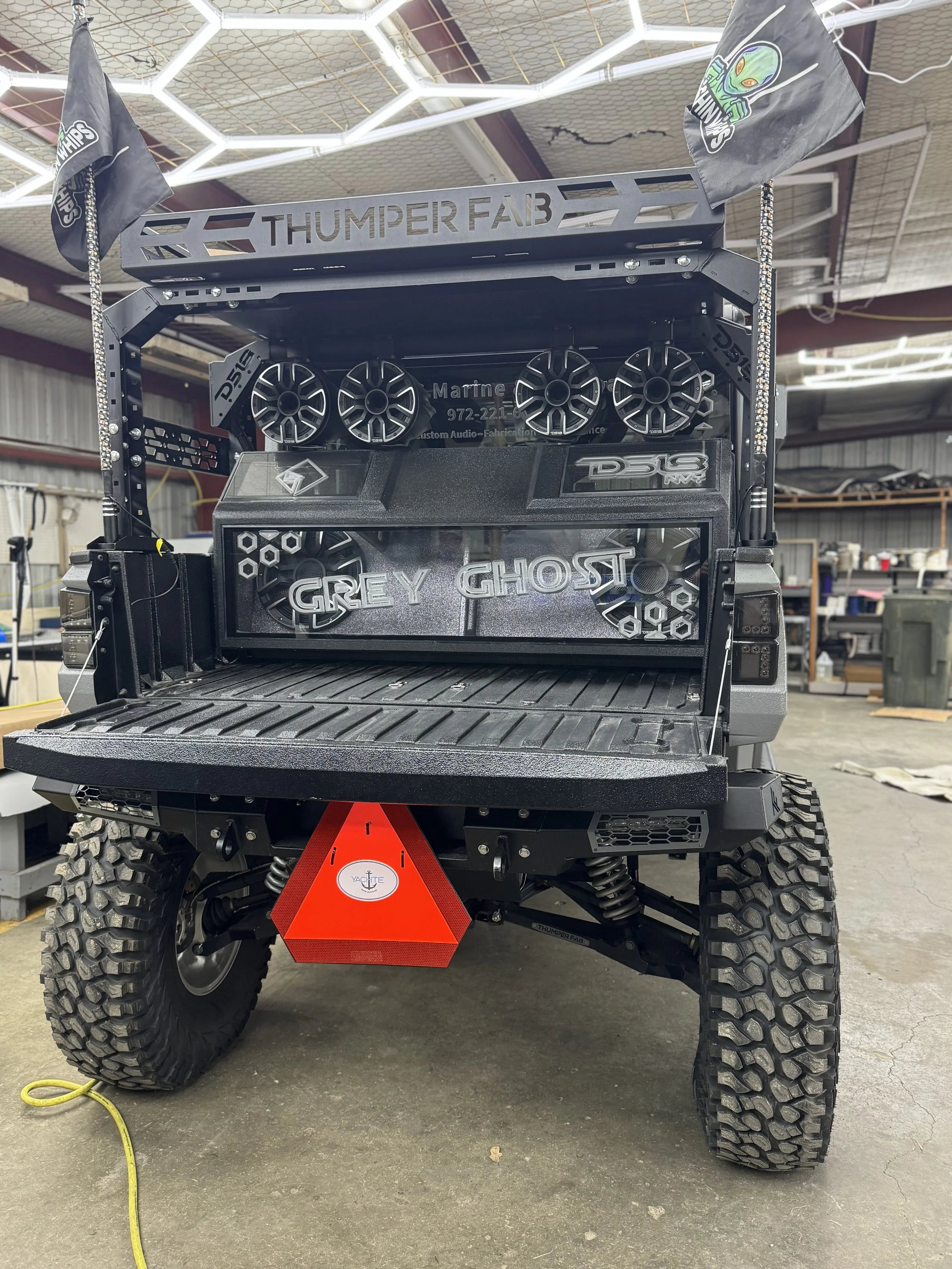 The back of a modified utility vehicle with large off-road tires, custom audio equipment, and a decorative panel that says 'Grey Ghost.'