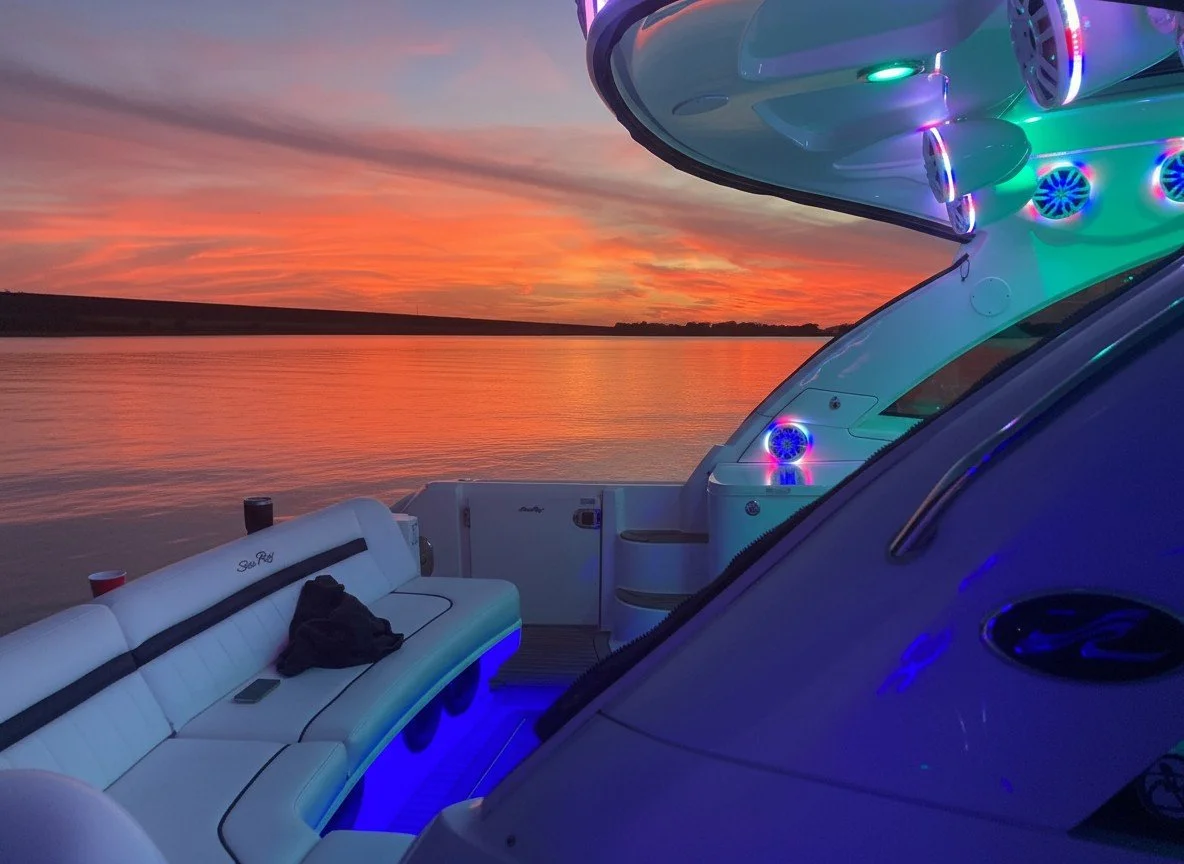 The image shows a white boat with lounge seating on a calm water body during a colorful sunset. The boat is illuminated with multicolored LED lights.