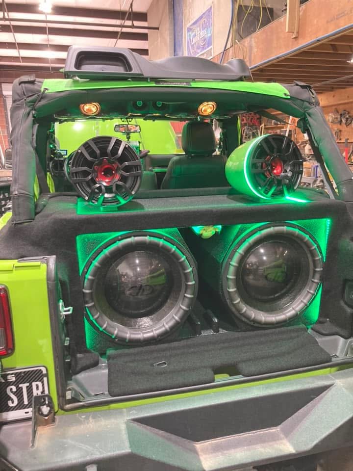 Off-road vehicle with multiple large, colorful speakers mounted on the roll cage, driving on a dusty trail in a mountainous area at sunset.