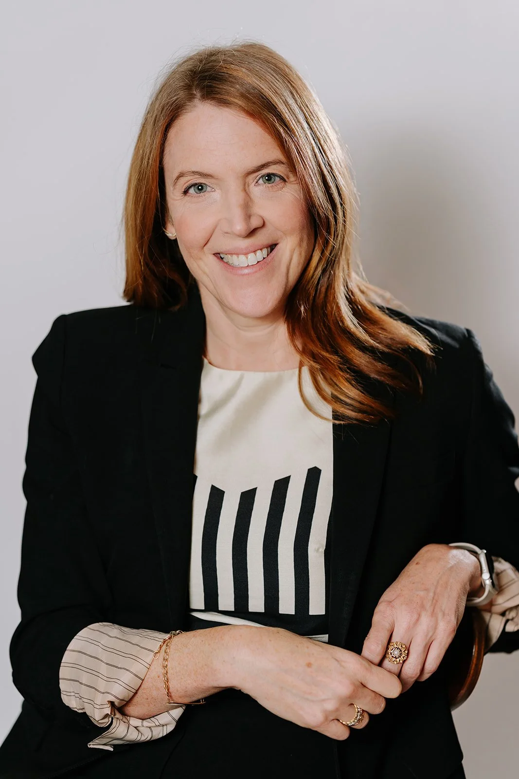 A woman with shoulder-length red hair and fair skin, smiling, wearing a black blazer over a white shirt with black stripe pattern, sitting with her arms crossed on her lap, on a light background.