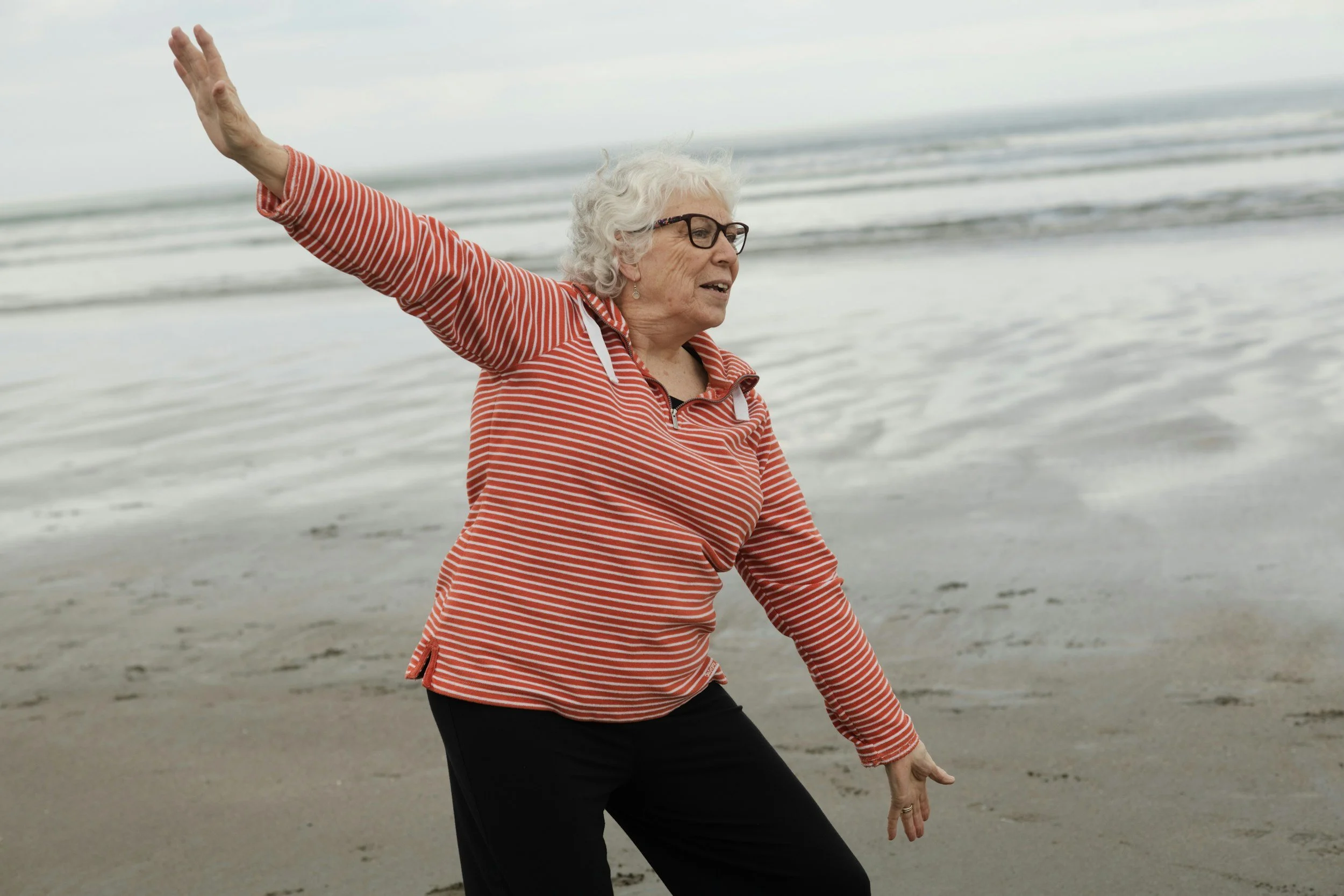 Older woman on the beach practicing yoga or stretching, wearing a red and white striped hoodie and black pants.