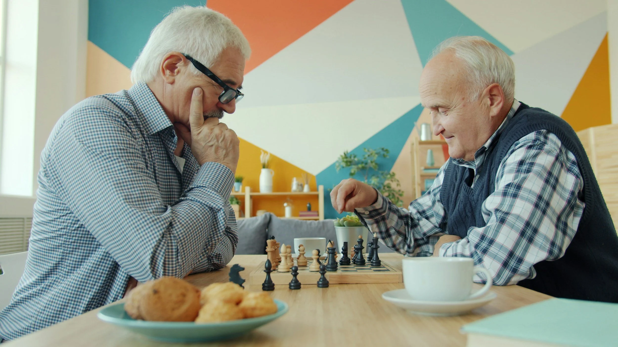 Two elderly men playing chess at a table, with a plate of cookies and cups, in a cozy room with colorful geometric wall art.
