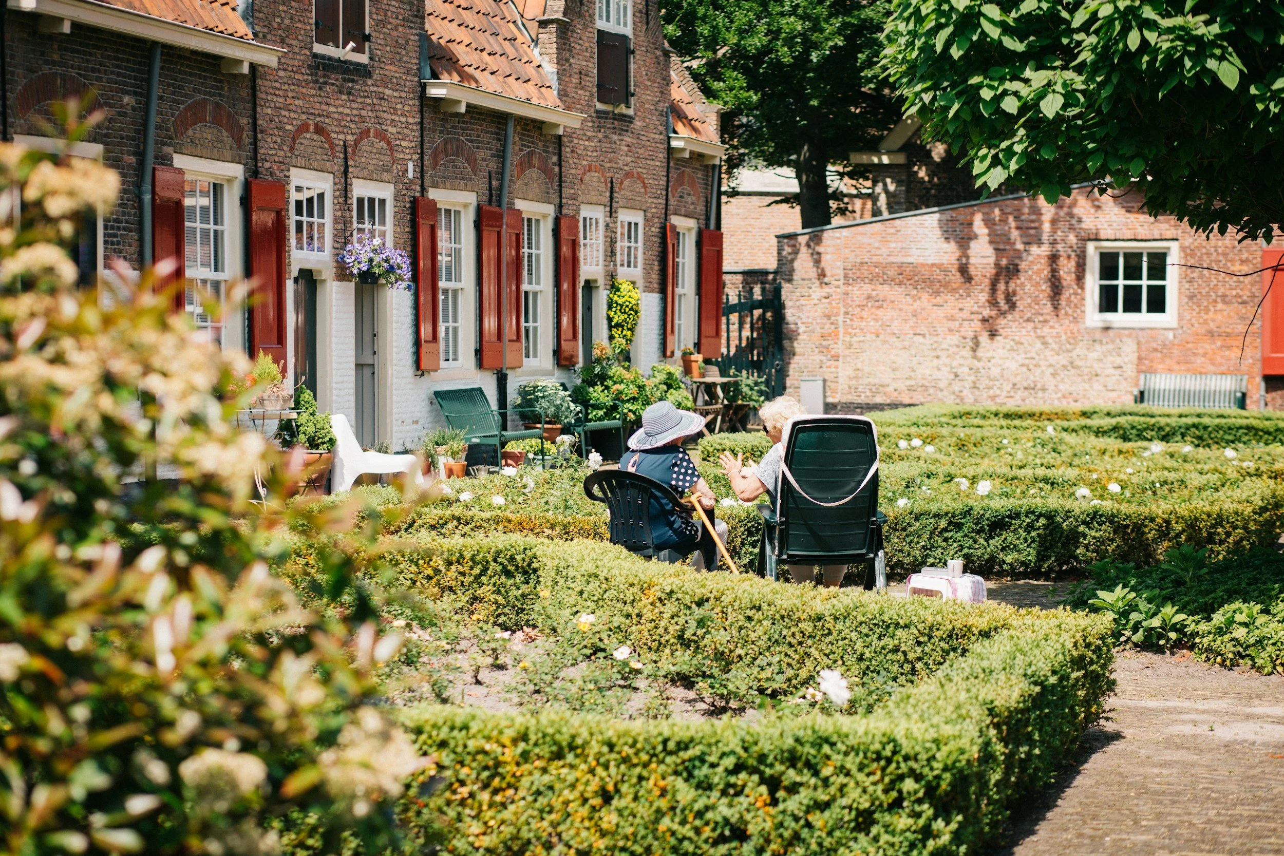 Two elderly women sitting and talking in a lush, green garden outside a charming brick building with red shutters, surrounded by potted plants and flowers, on a sunny day.