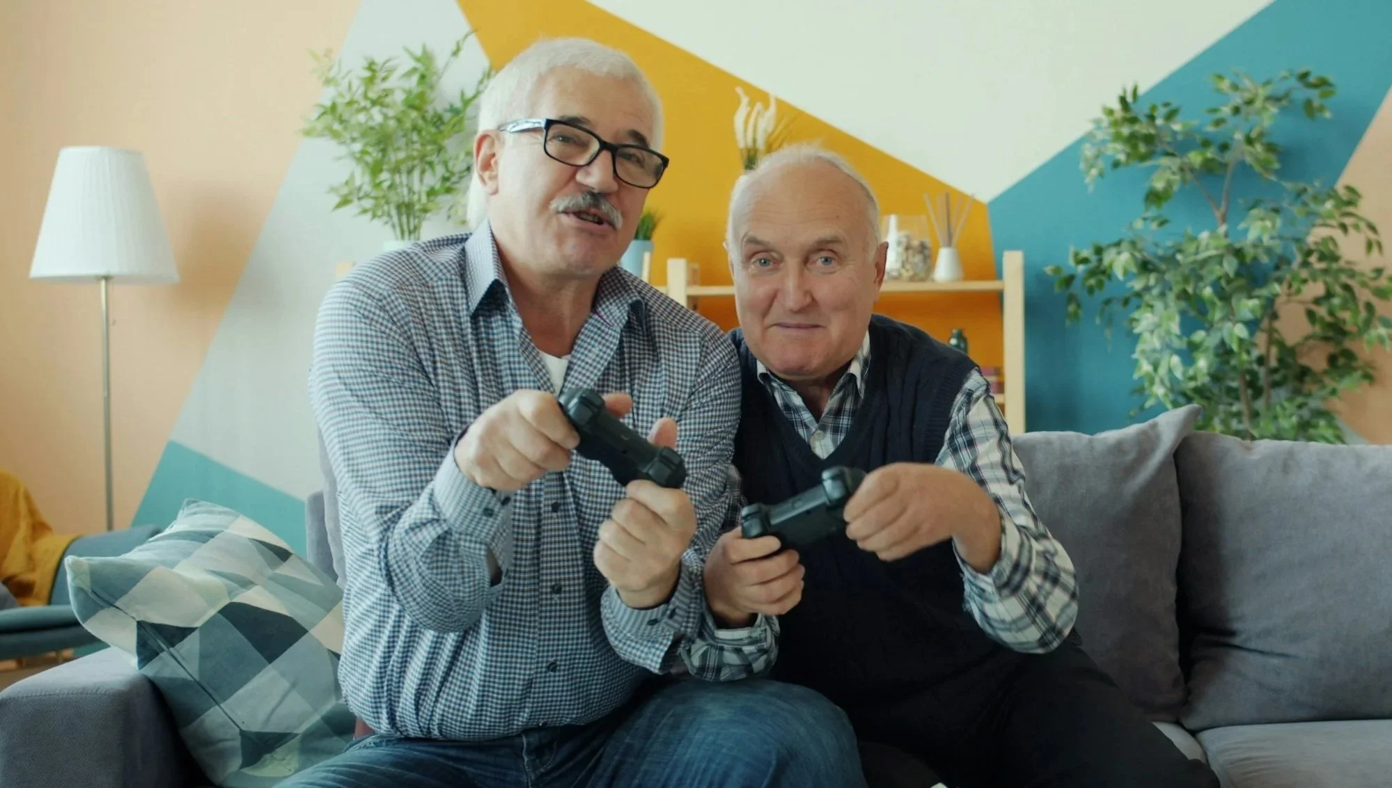 Two older men with gray hair and wearing plaid shirts, sitting on a gray sofa, playing video games with black controllers. They are in a colorful living room with plants, a lamp, and decorative wall art.