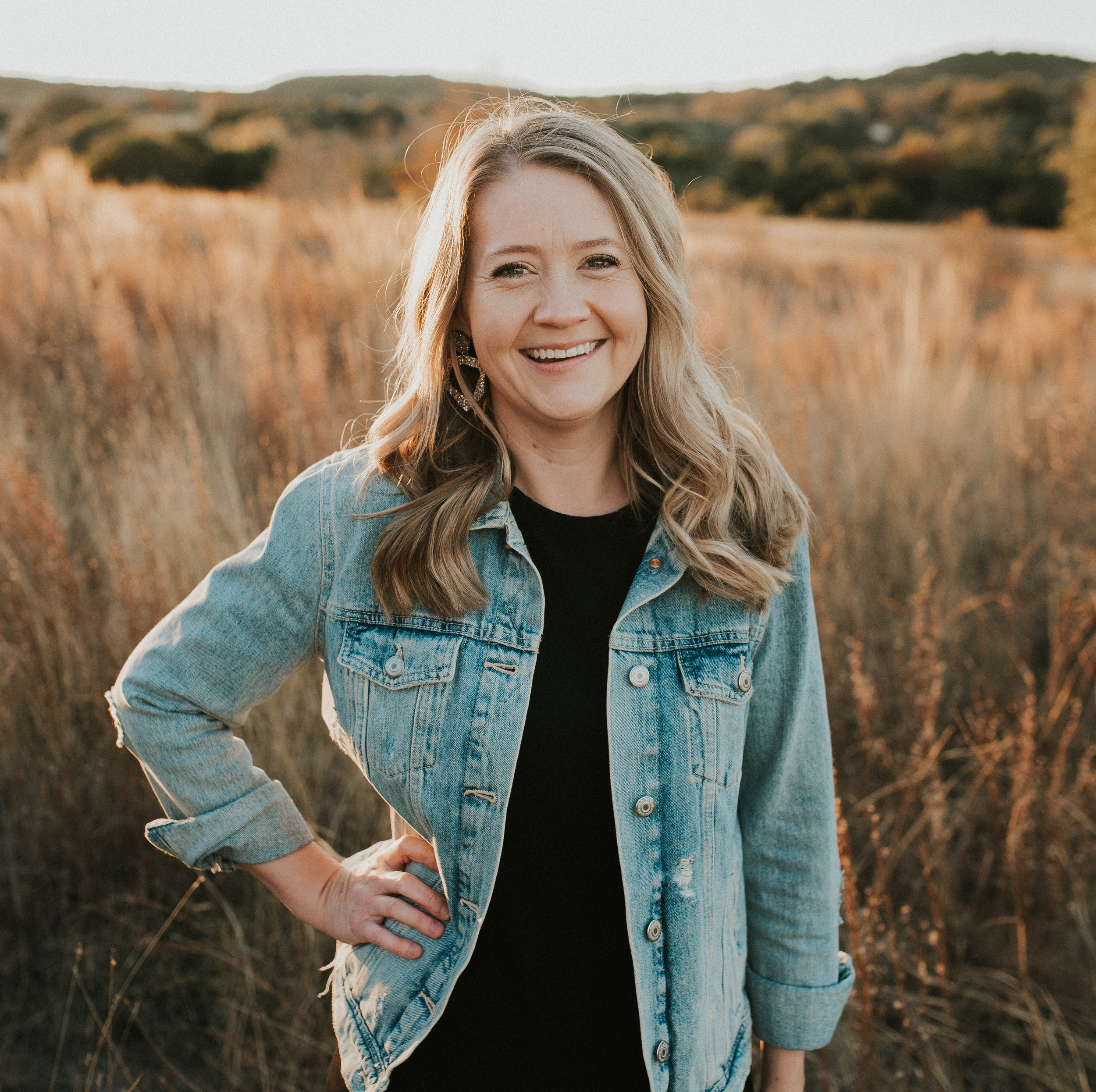 A woman with blonde hair smiling outdoors in a field during sunset, wearing a denim jacket and black shirt.
