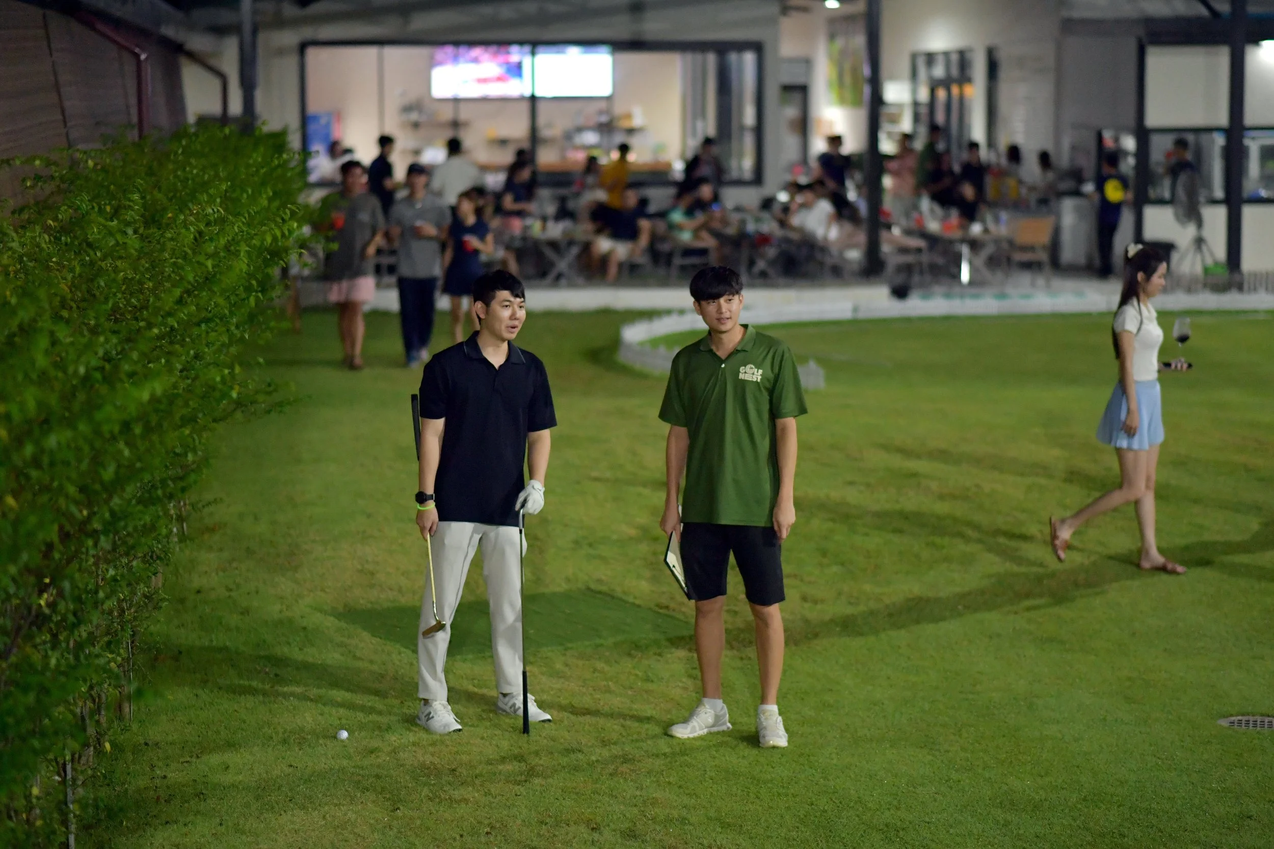 Two young men standing on a golf course, one holding a golf club, with a woman walking by in the background holding a glass of red wine. In the background, people are dining inside a building.
