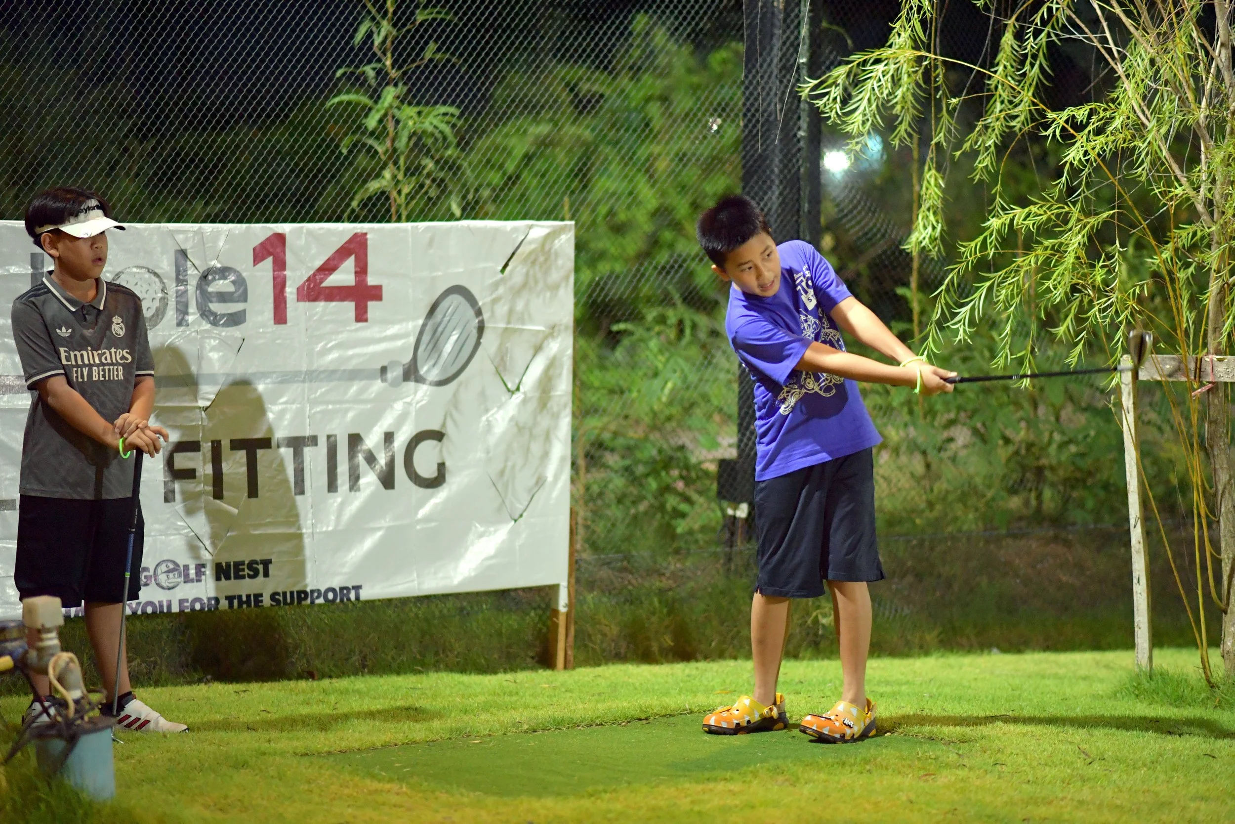 Two boys playing golf on a small outdoor practice area at night. One boy in a blue shirt is swinging a golf club, while the other boy in a black Adidas shirt and visor watches. There is a large sign with a golf ball graphic and the words 'Golf 14 FIT