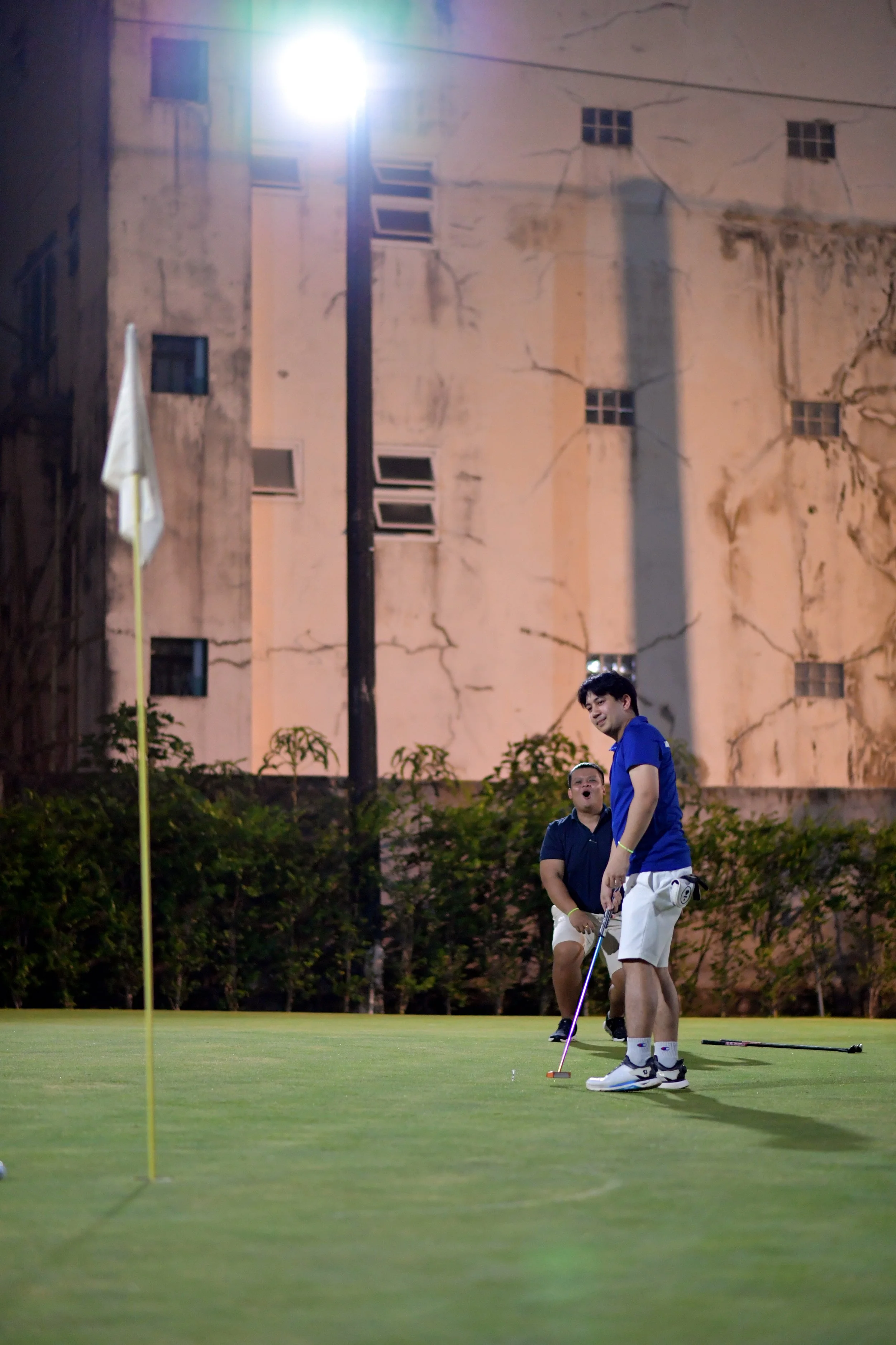 Two men playing golf on a green at night, with a large, cracked building and a tall streetlight in the background.
