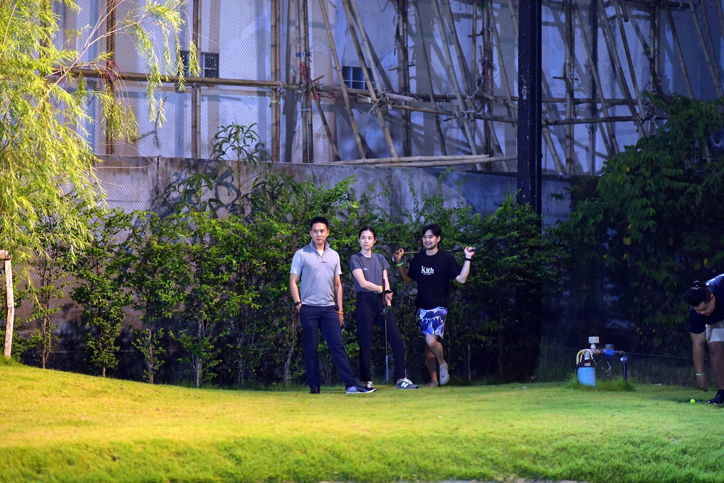 Three people standing on a well-lit grassy area at night, with trees and a construction site in the background. One person is holding a golf club, another has their hands in their pockets, and the third is squatting near a blue container.
