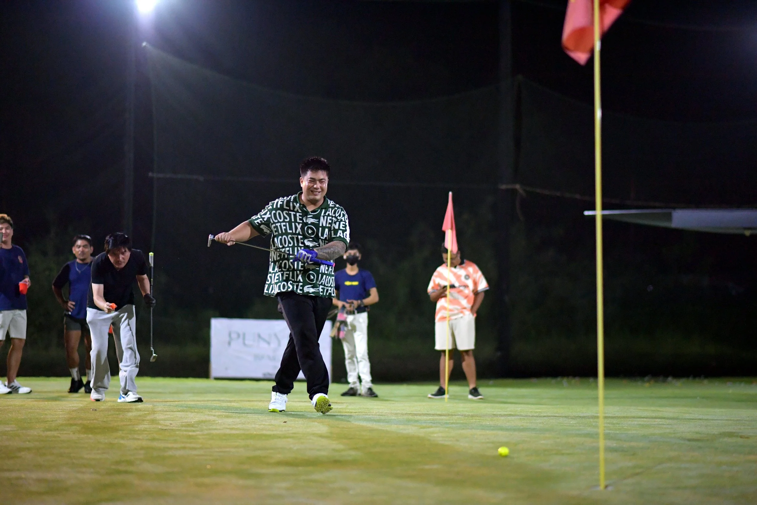 Group of people playing golf on a putting green at night. One person is preparing to putt, while others stand and watch. The scene is lit by artificial lights, with flags marking the holes in the background.