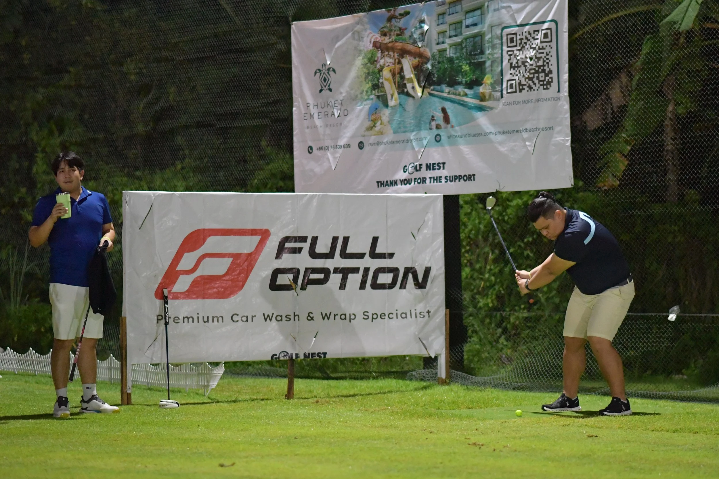 Two men on a golf course, one preparing to hit a golf ball with a club while the other stands nearby holding a drink, with advertising banners in the background.