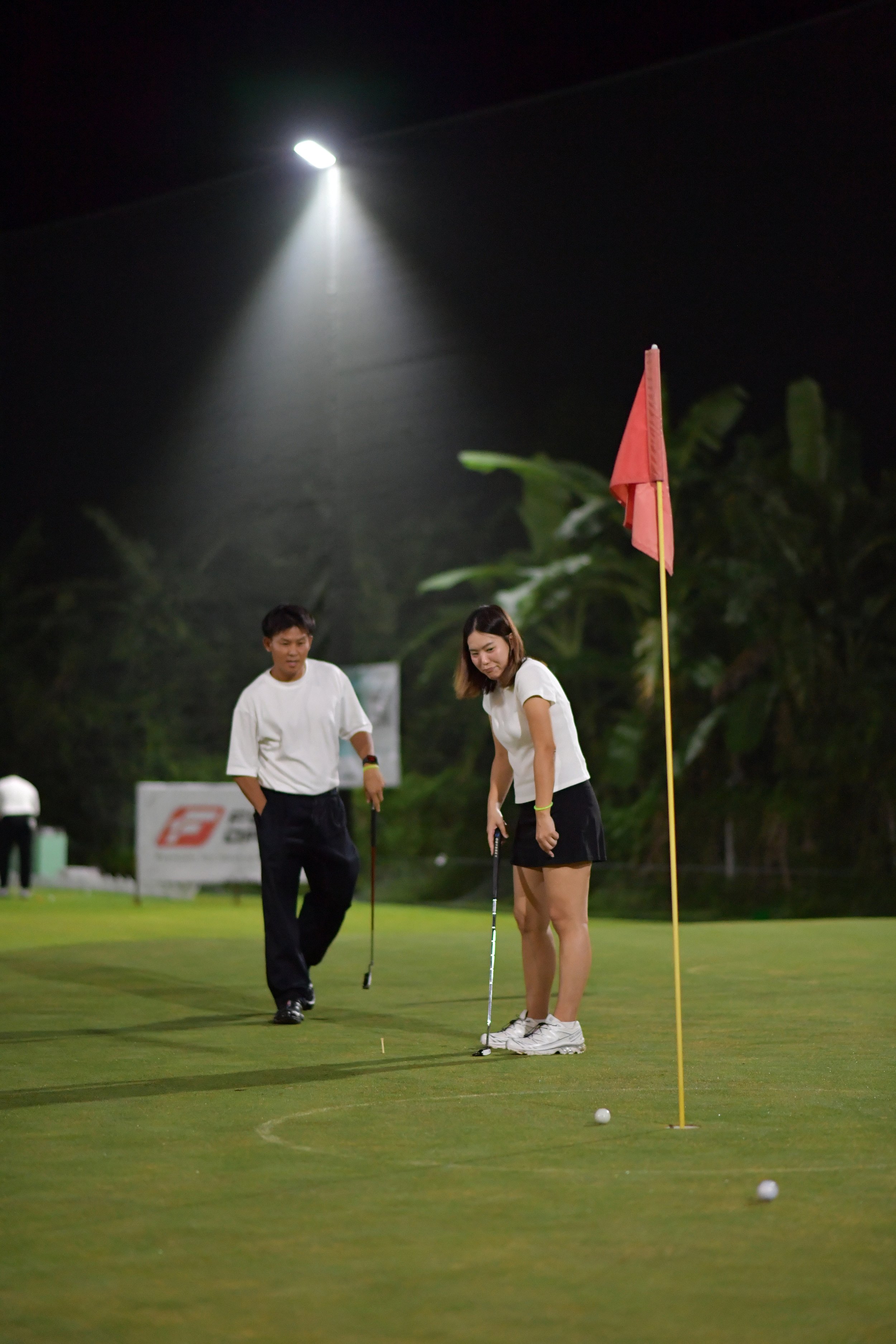 Two people practicing putts on a golf green at night, with a flagstick and golf balls nearby, under artificial lighting.