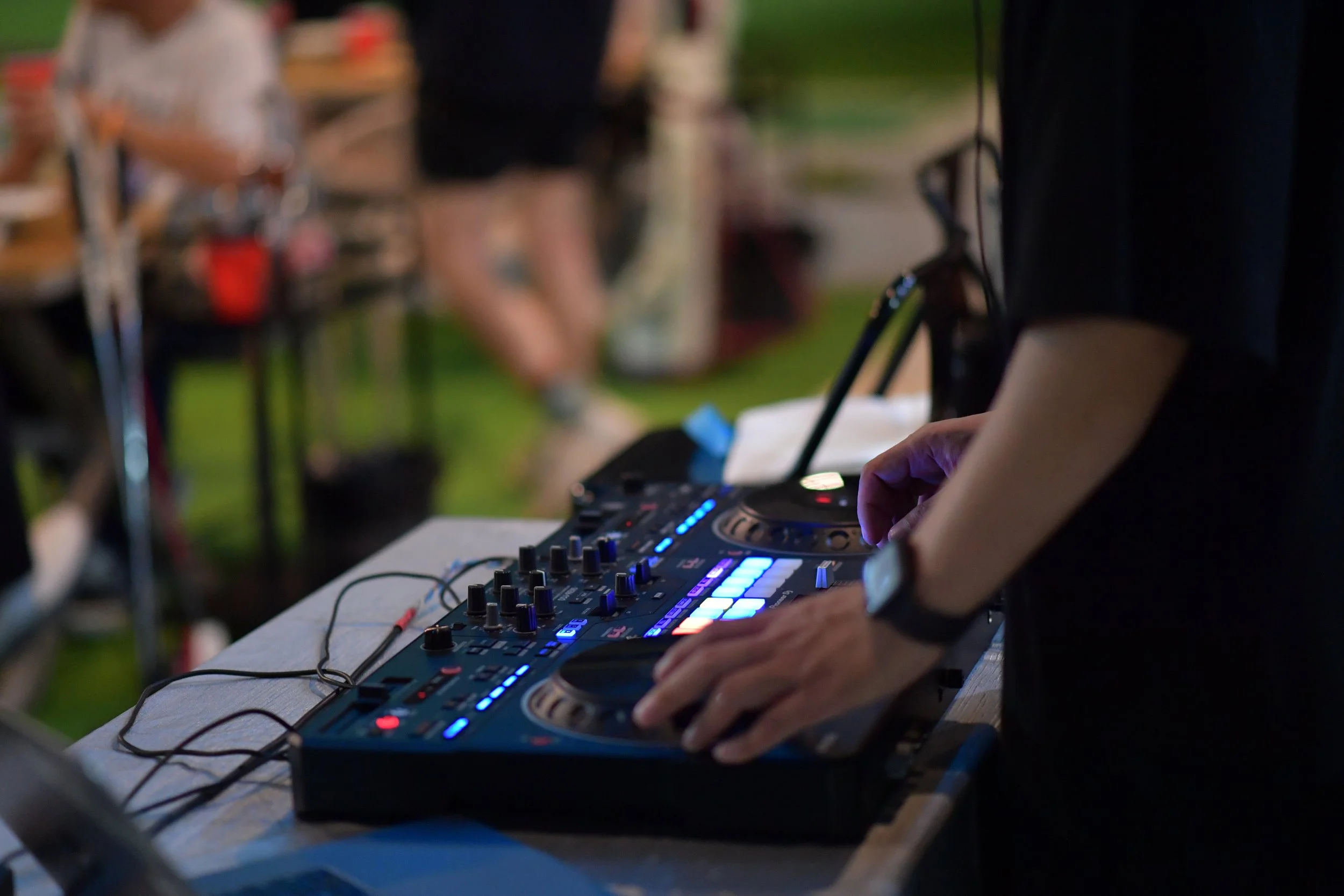Close-up of a DJ adjusting controls on a DJ mixer with colorful LED lights at an outdoor event, with blurred people in the background.