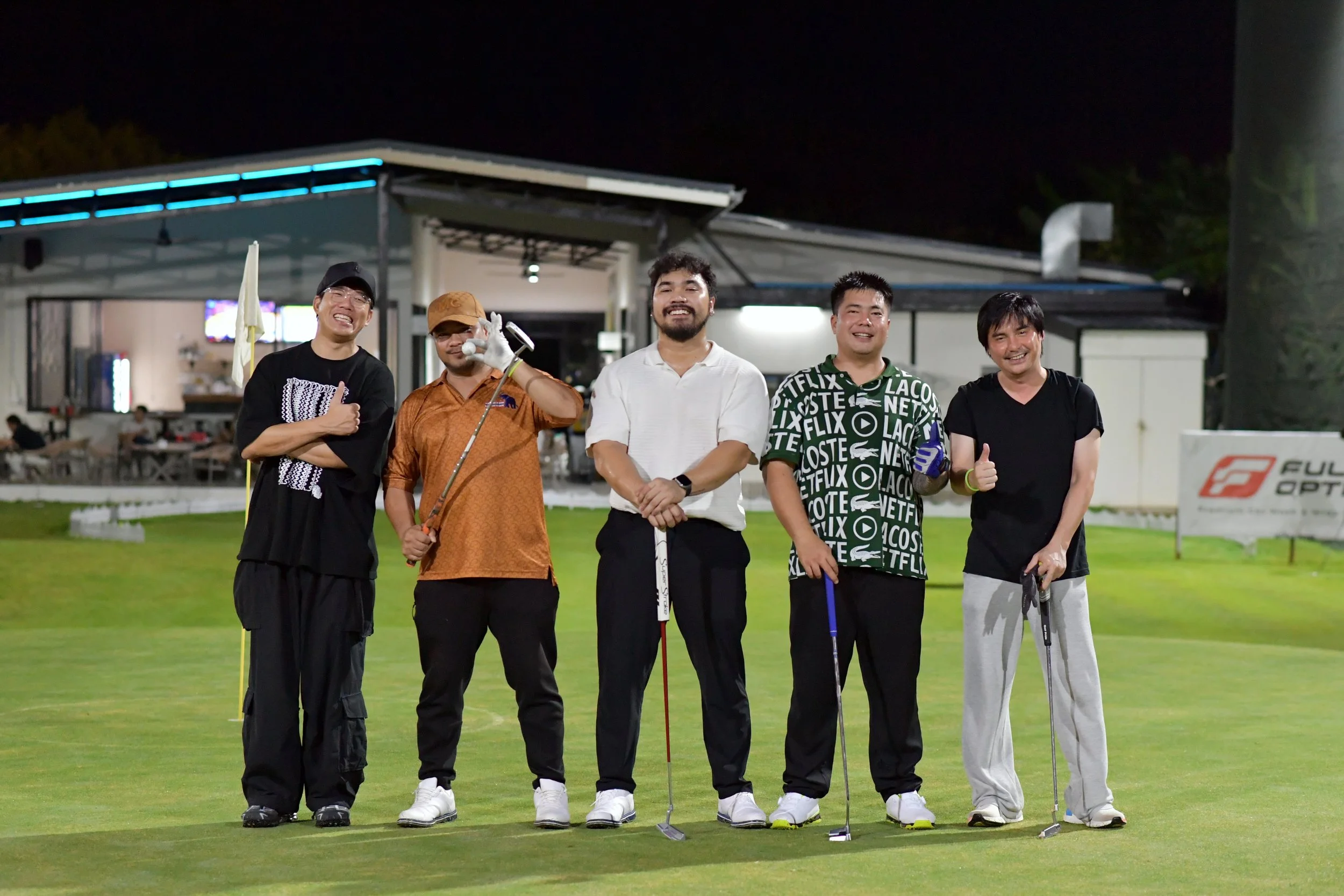 Five men standing on a golf course at night, smiling and posing for a photo, each holding a golf club, with a clubhouse in the background.