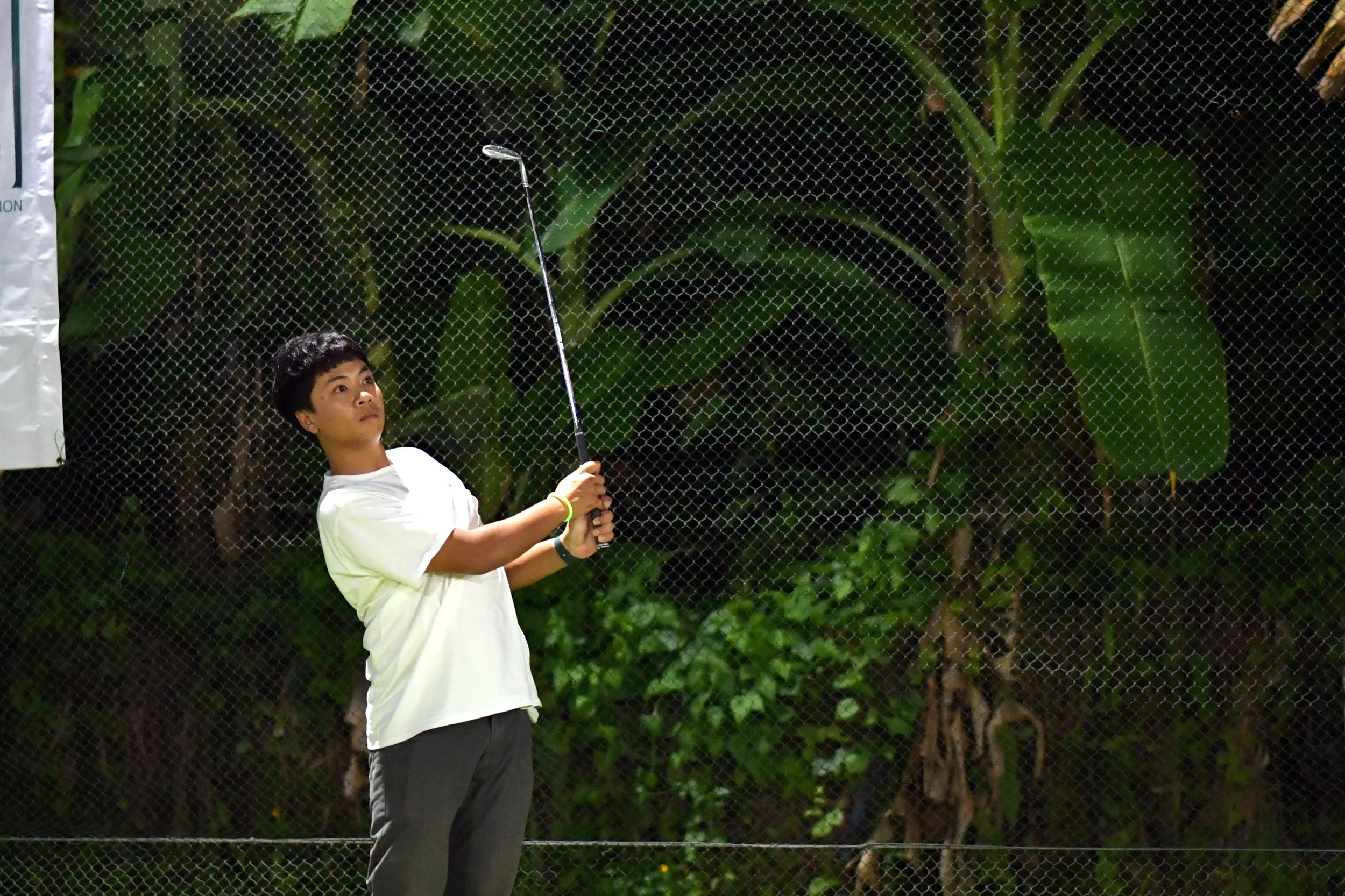 A young boy playing golf on a night golf course, swinging his club near a net with greenery in the background.