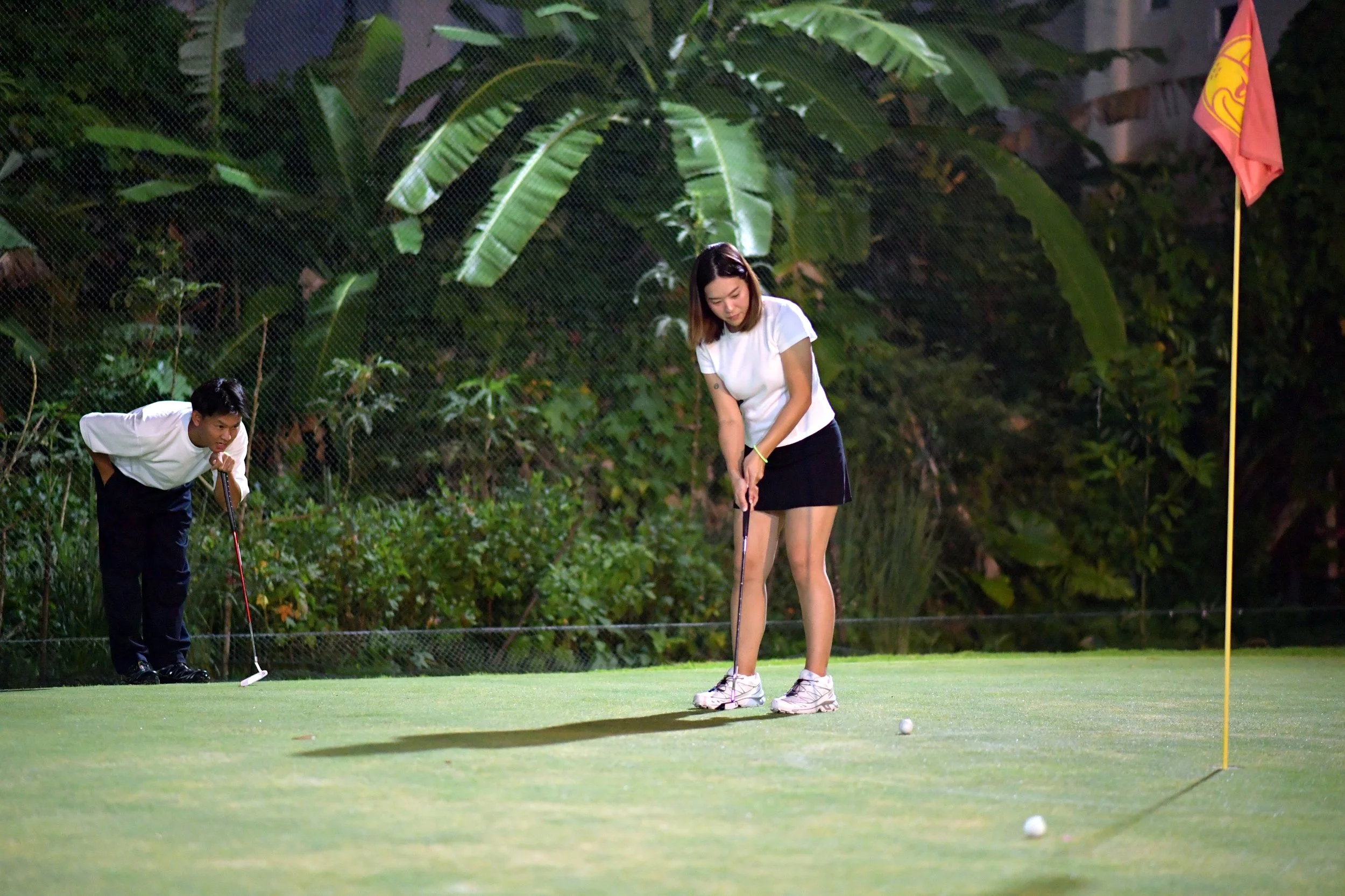 A woman practicing golf on the putting green with a man observing at night.