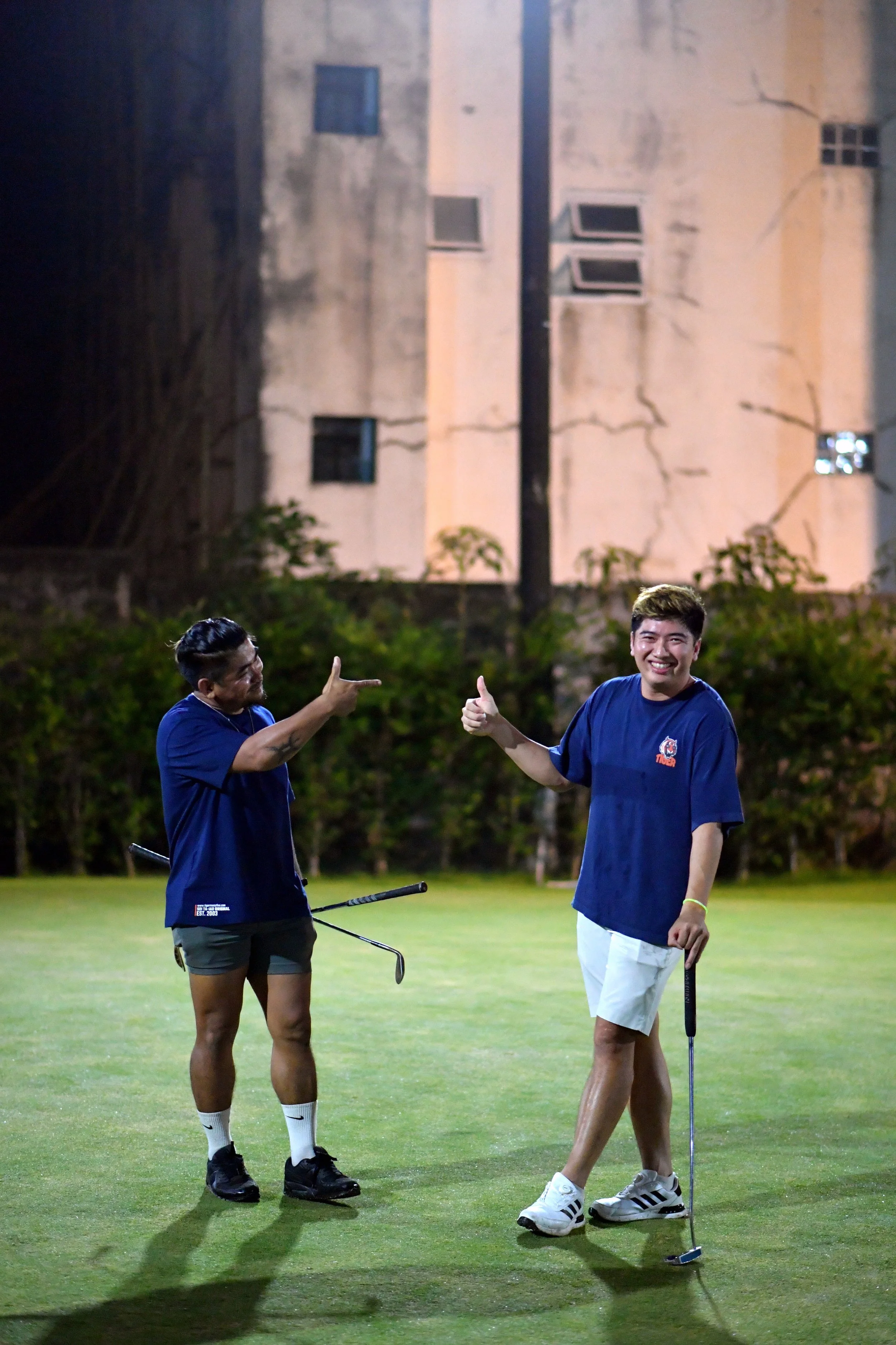Two men standing on a golf course at night, smiling and giving thumbs-ups, one holding a golf club.