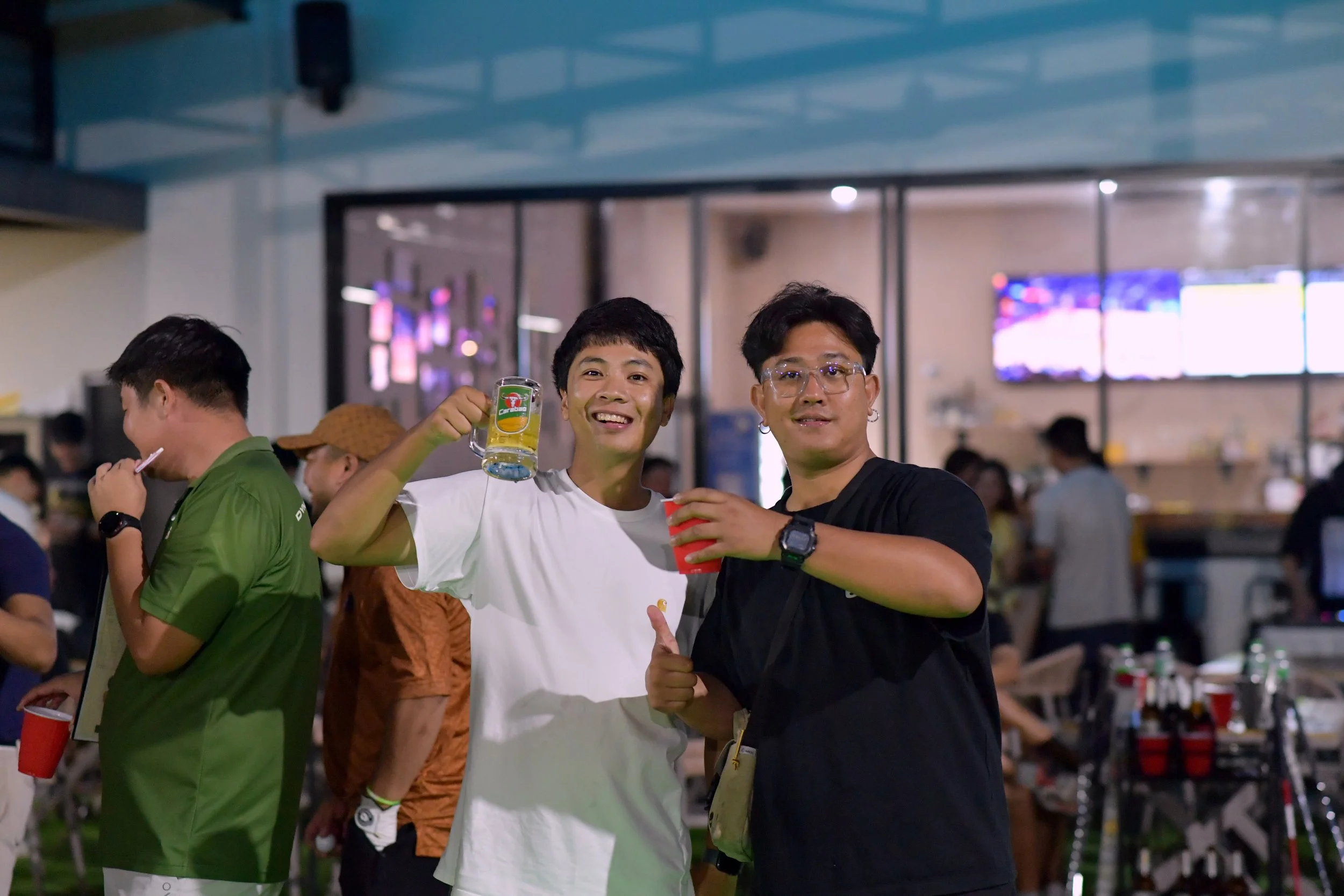 Two young men enjoying drinks and celebrating, standing indoors at a social gathering or party, with other people in the background.