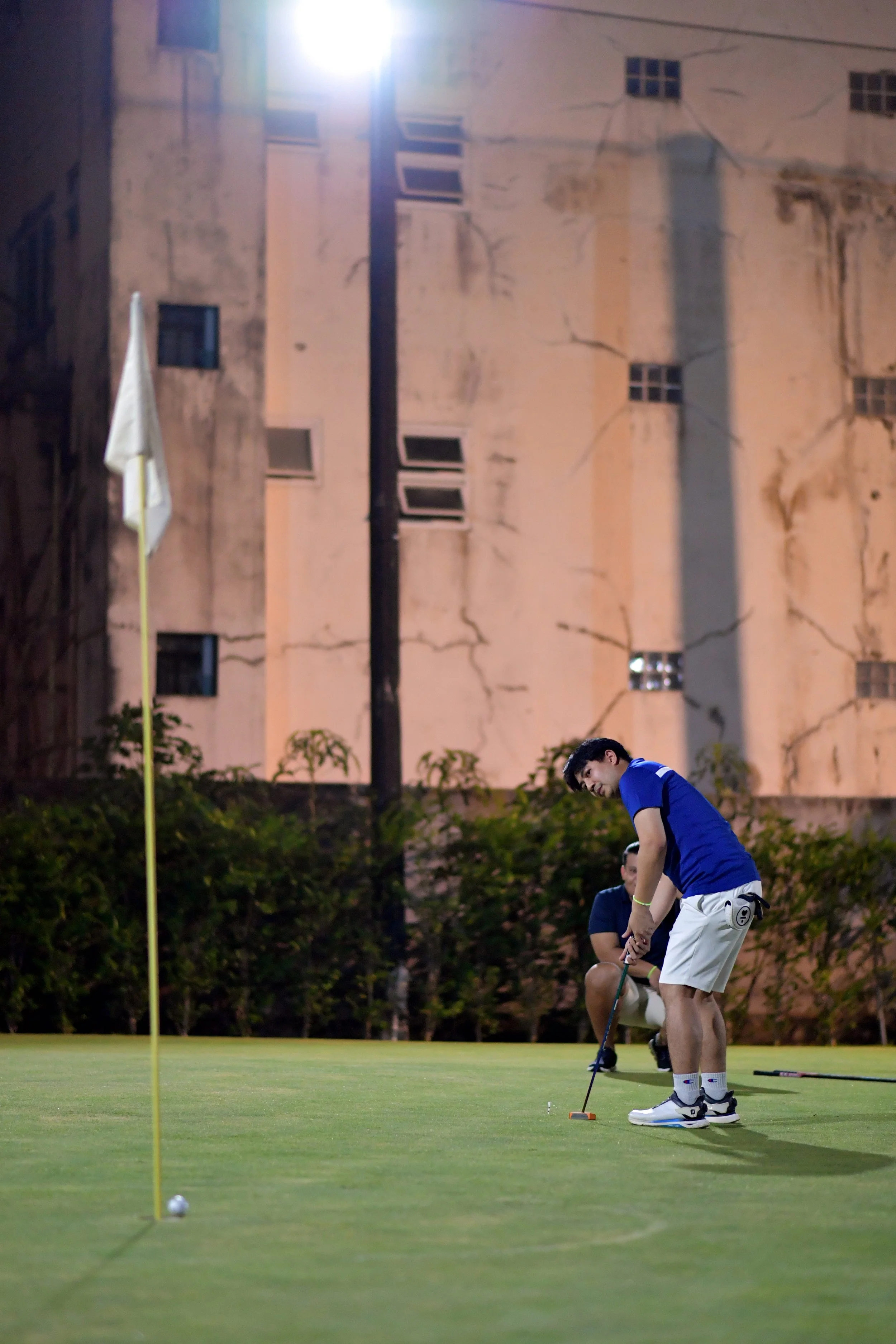 Two young men playing golf on a putting green at night, with a tall, weathered building in the background, illuminated by a nearby light.