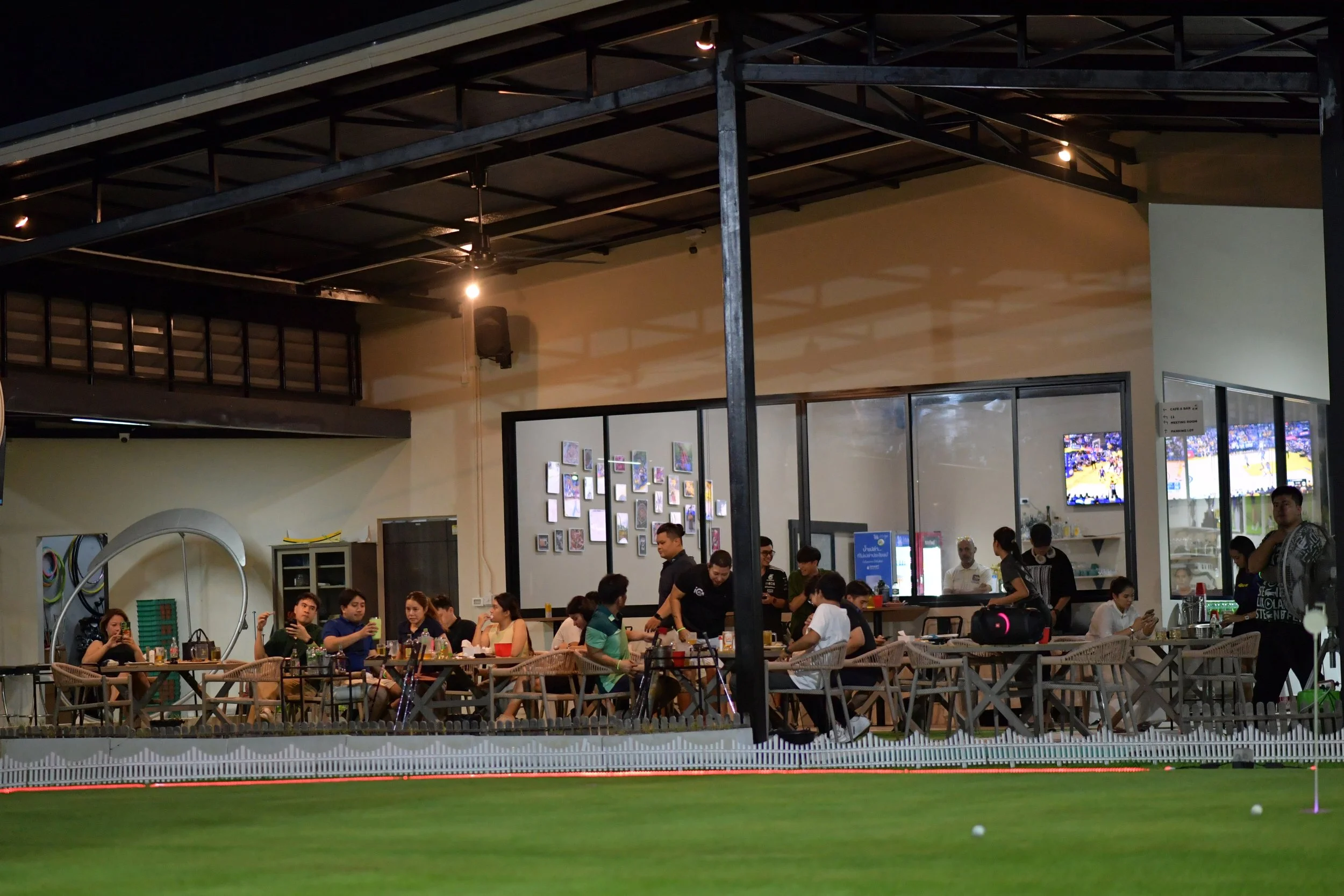 Indoor dining area with people seated at tables, some using mobile phones, behind a house or club with a mini golf course in the foreground.