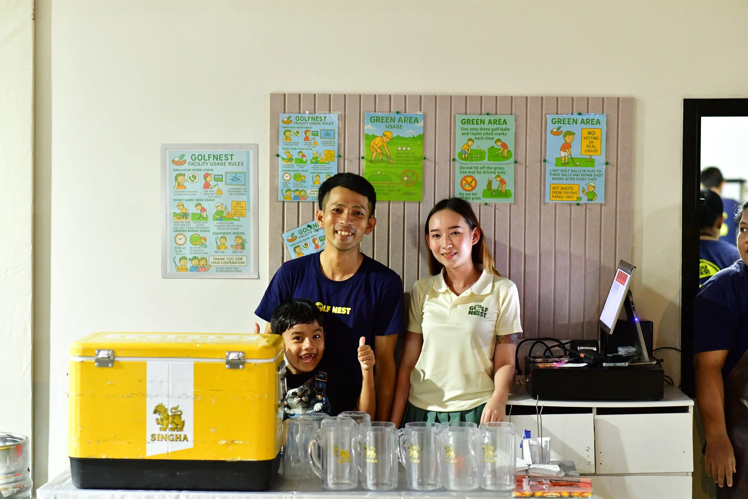 A group of three people, a man, a woman, and a boy, standing behind a table with a yellow cooler and several glass mugs, smiling at the camera. Behind them are signs with rules and guidelines for a golf facility.