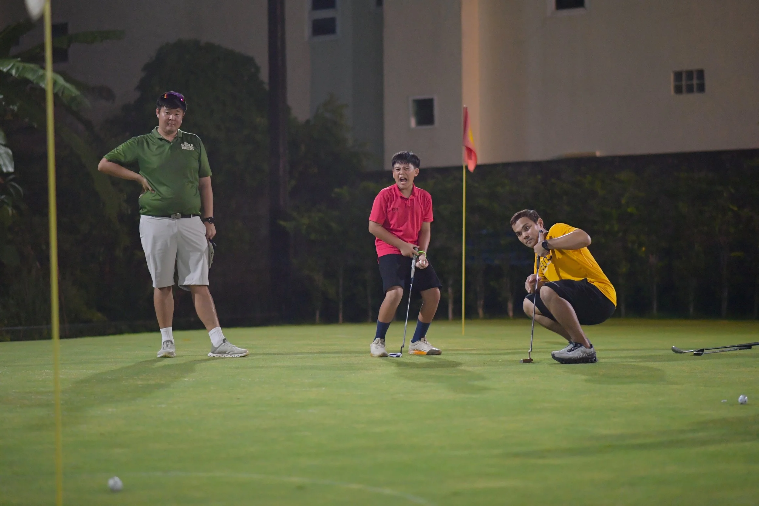 Three people on a golf course at night, two crouching and one standing, with golf clubs and a golf ball near the hole, possibly engaged in a game or practice.