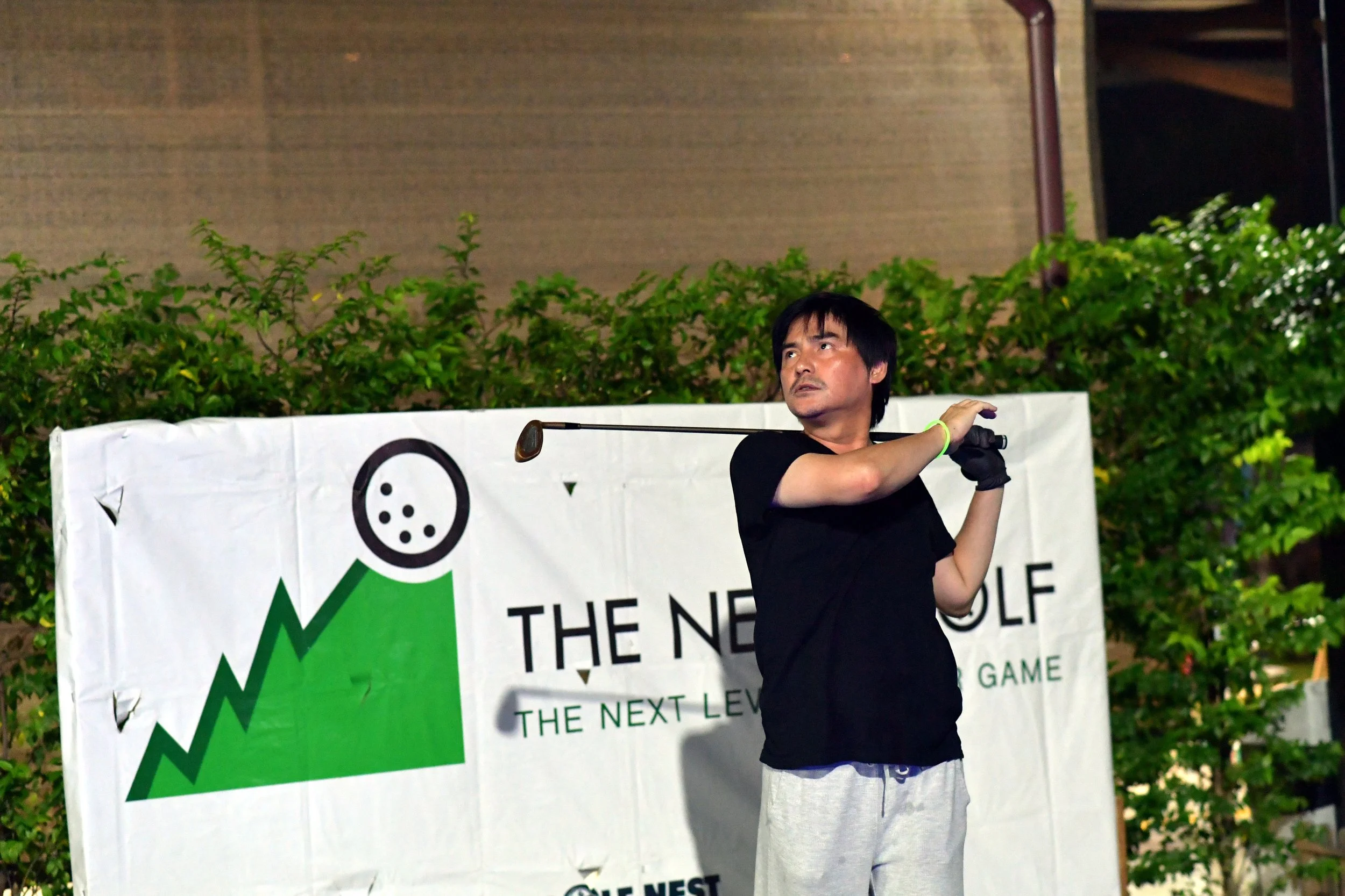 A man in black shirt and light-colored pants holding a golf club on a green and white banner that reads 'The Nest Golf,' with bushes and building in the background.