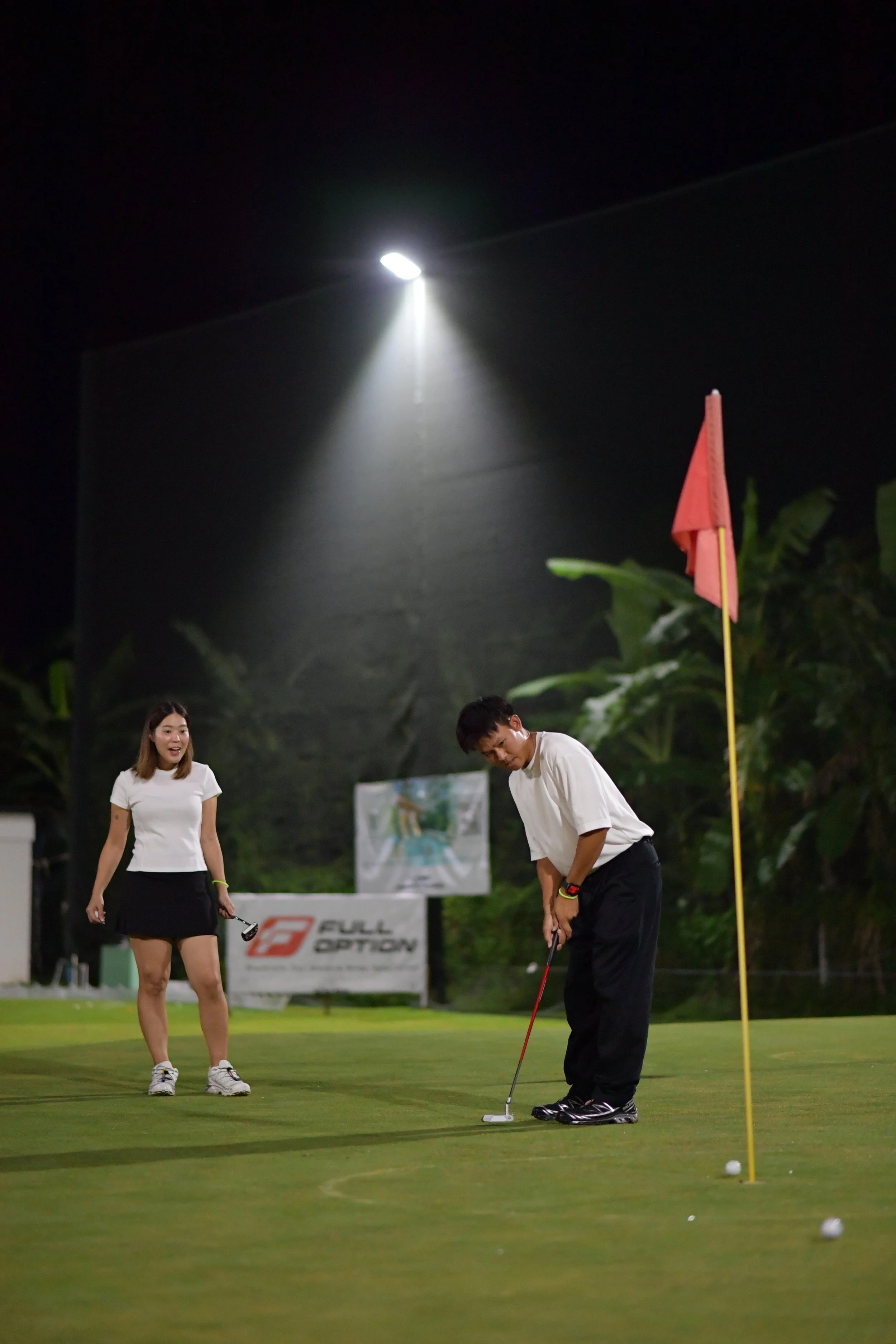 Two people practicing golf on a green at night, with one person putting and the other watching. There is a red flag on a pole and golf balls on the ground.