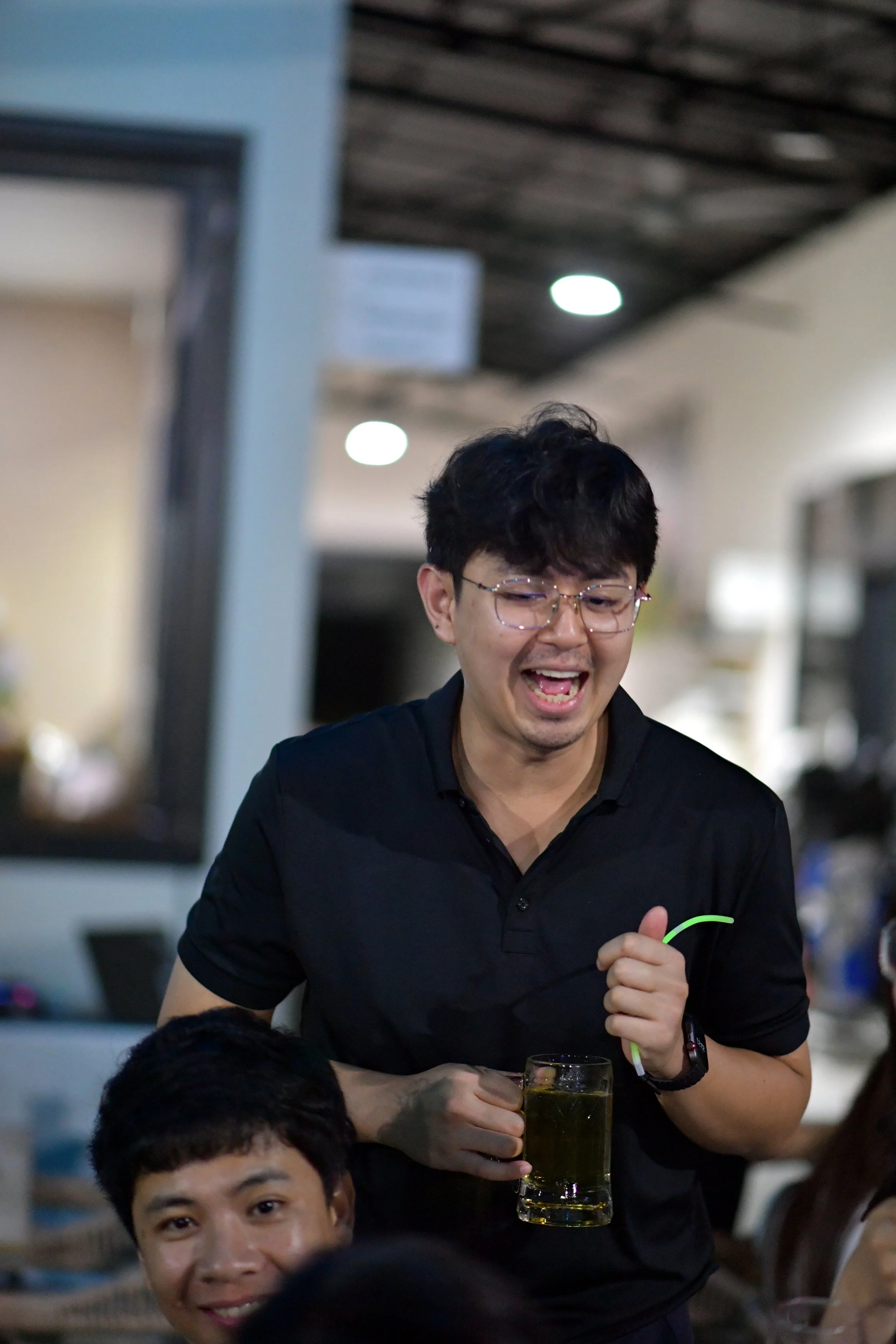 A young man with glasses and black hair, wearing a black shirt, is smiling and holding a glass mug filled with greenish beverage. Another person, a young man with short dark hair, is partially visible in the foreground, smiling at the camera. The bac
