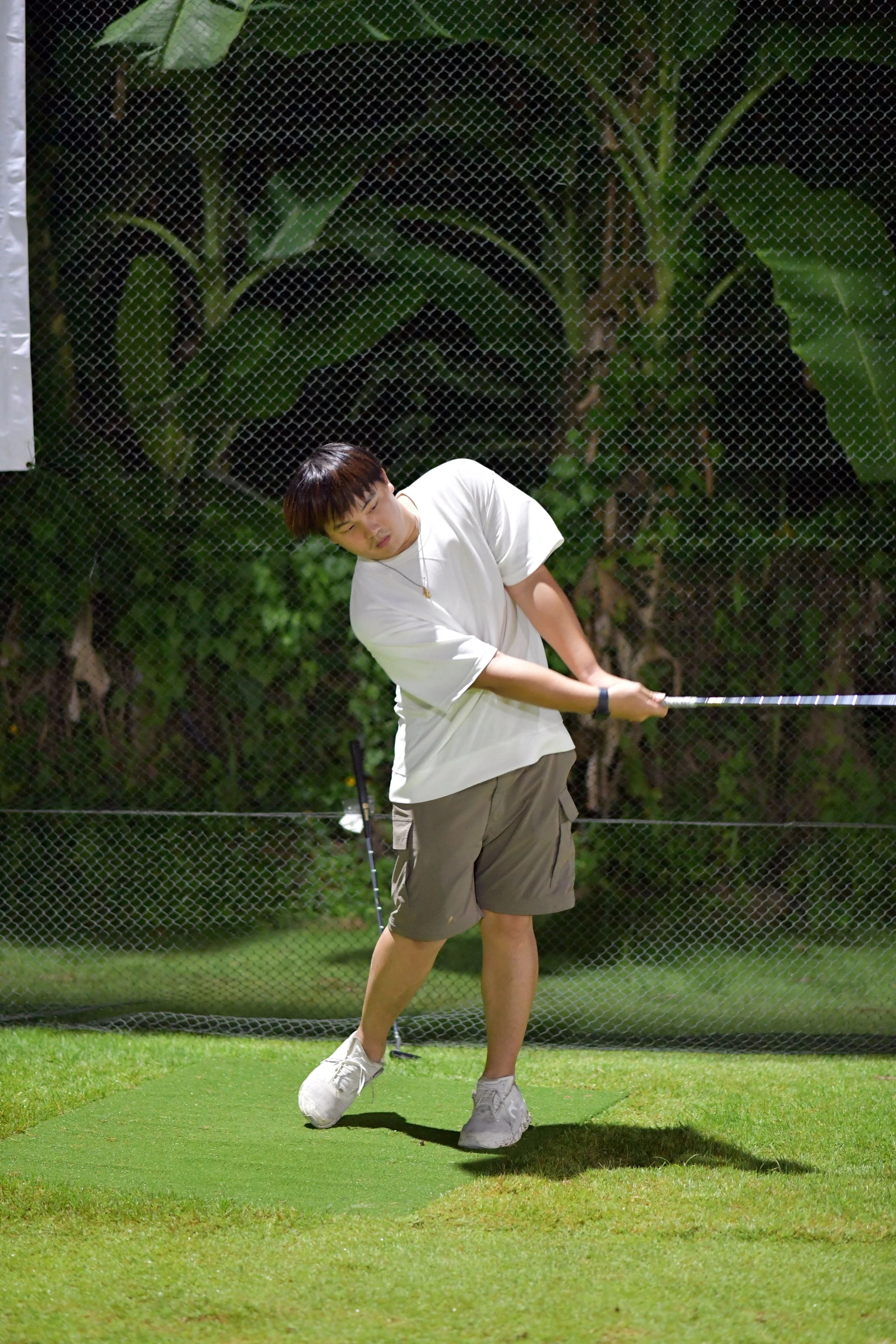 A young man is playing golf, swinging a club on a grassy area, with a mesh fence and large green leafy plants in the background.