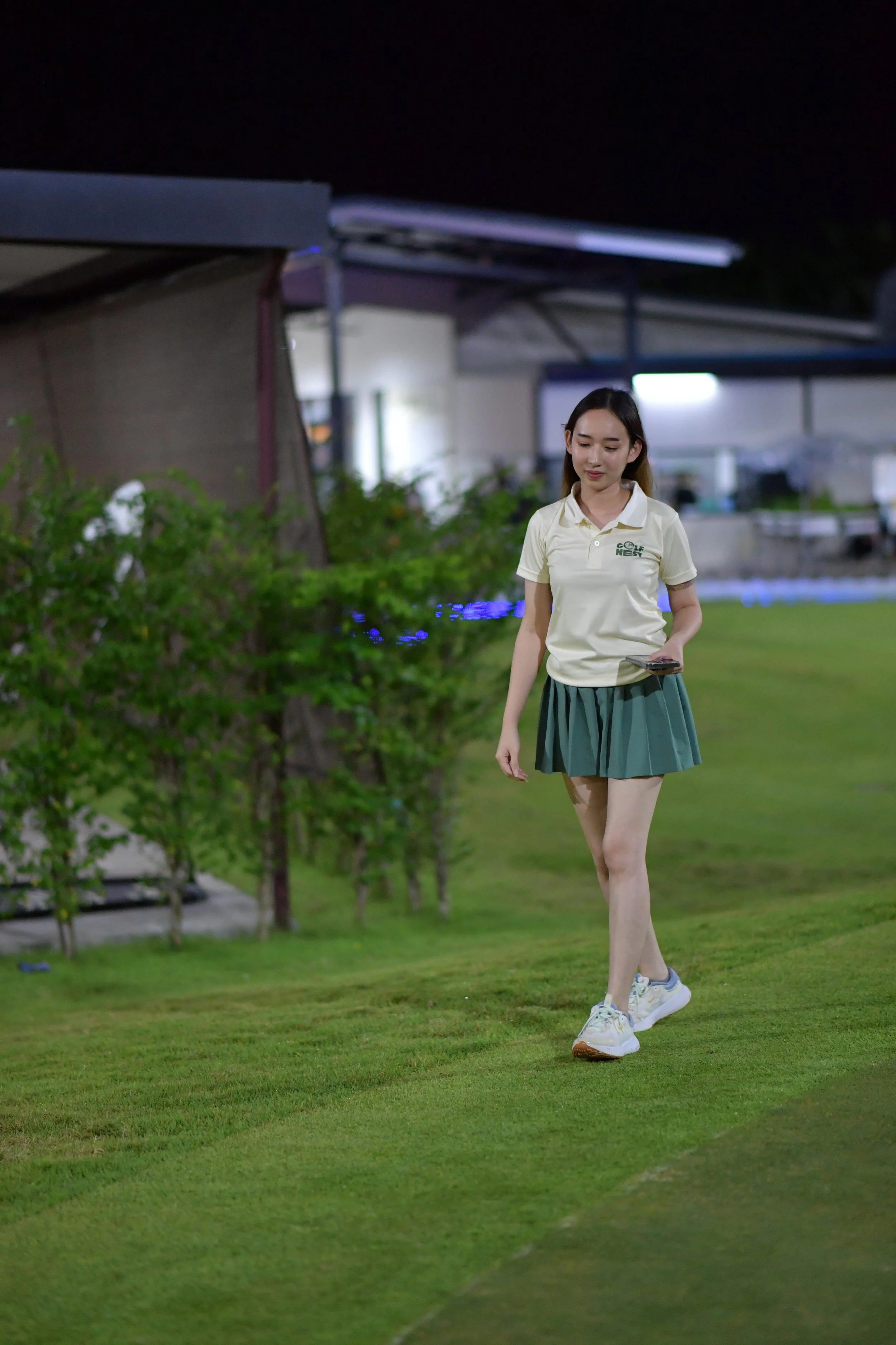 Young woman walking on a grassy field at night, holding a smartphone, wearing a light yellow polo shirt and a green skirt.